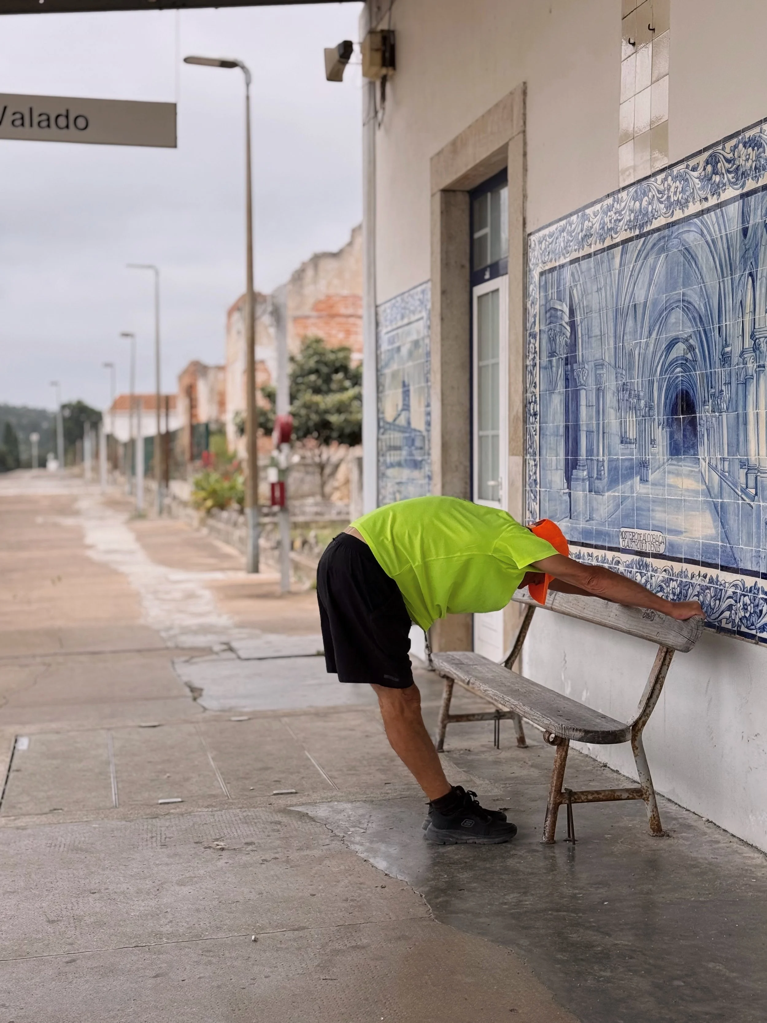 A man in a neon yellow shirt and black shorts bends over a wooden bench on a sidewalk, reaching towards it. Behind him, there is a white wall with large blue and white tile artwork depicting an arched interior scene, and a small window. The scene app