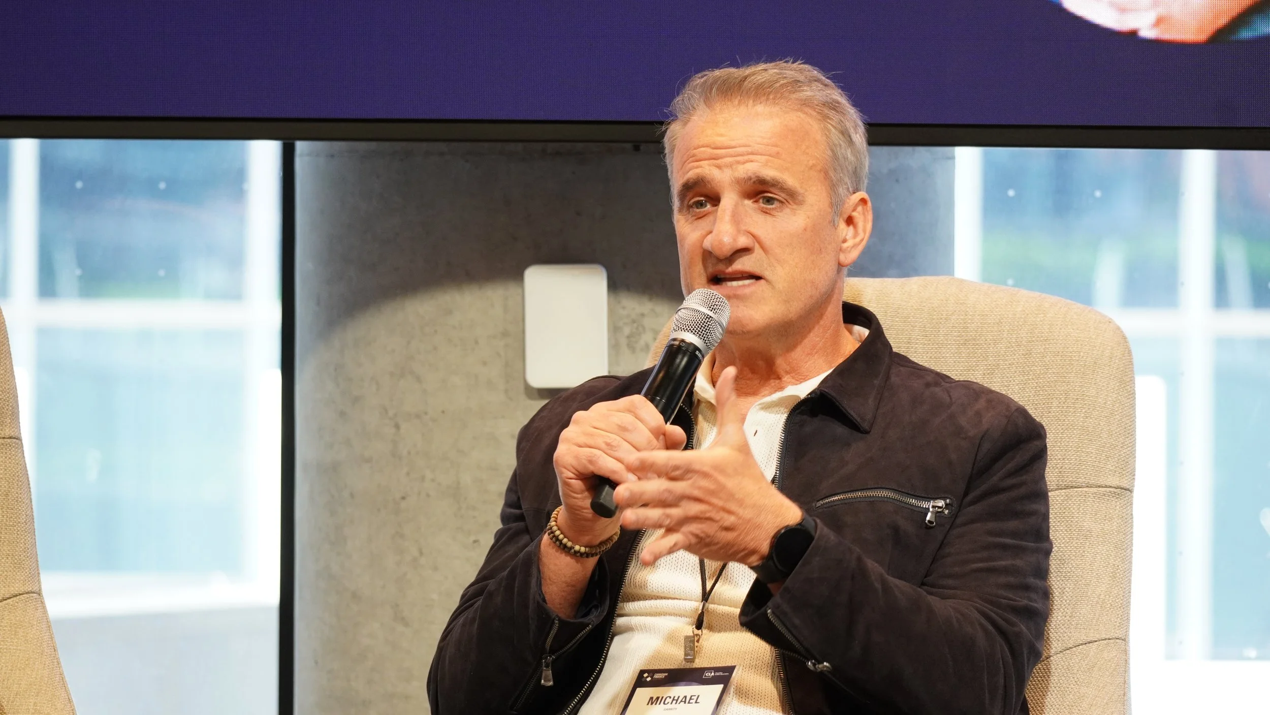 A middle-aged man with gray hair, wearing a black jacket, white shirt, and a smartwatch, speaking into a microphone during a panel discussion or interview.