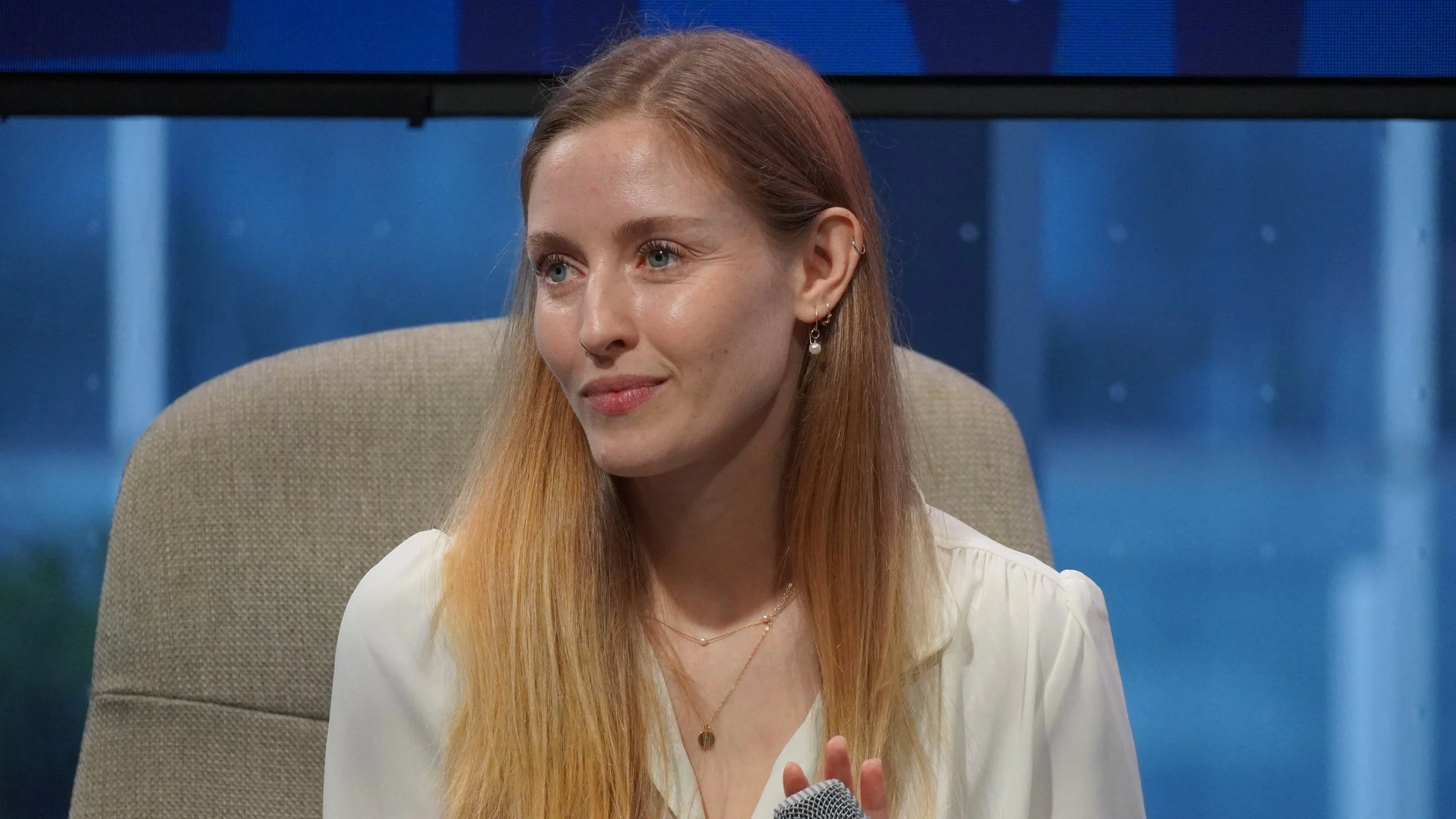 A woman with long reddish hair sitting on a beige chair in front of a dark blue background, holding a microphone in her hand, wearing a white blouse and jewelry.
