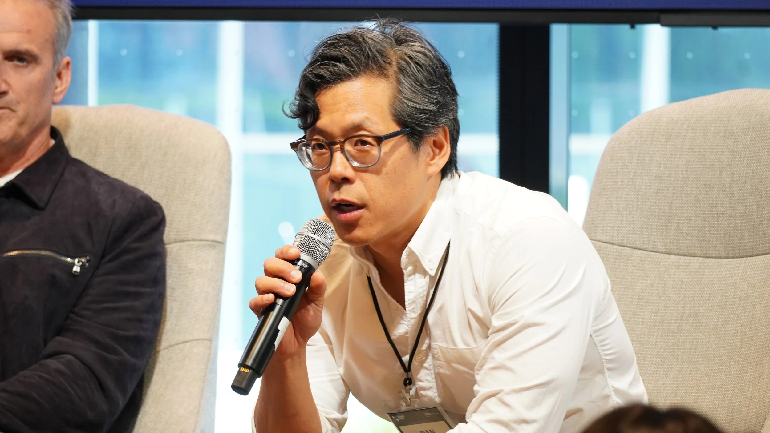 A man with glasses and a white shirt speaking into a handheld microphone during a panel discussion, seated in a beige chair with another person partially visible next to him.