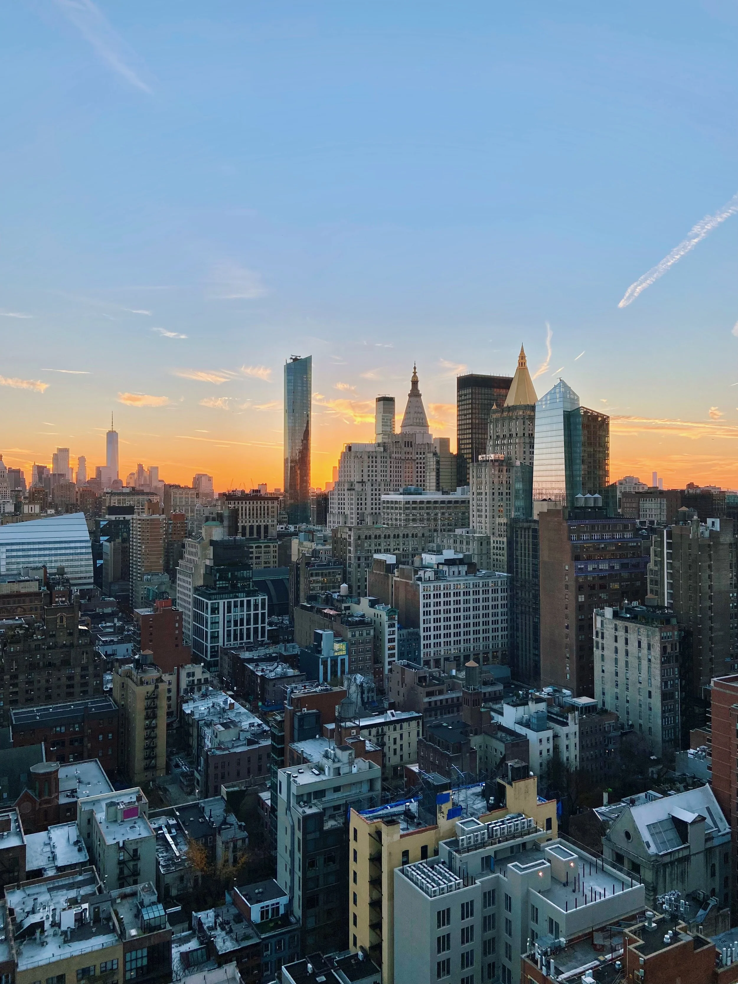 City skyline during sunset with tall skyscrapers, including One World Trade Center, and a blue sky with clouds.