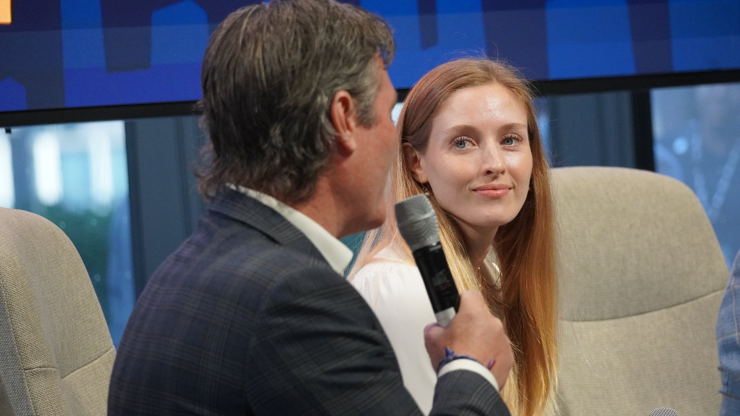 A man with gray hair holding a microphone and speaking, looking at a young woman with long red hair who is seated and listening with a slight smile. They are in a conference or panel setting with beige chairs and large windows in the background.