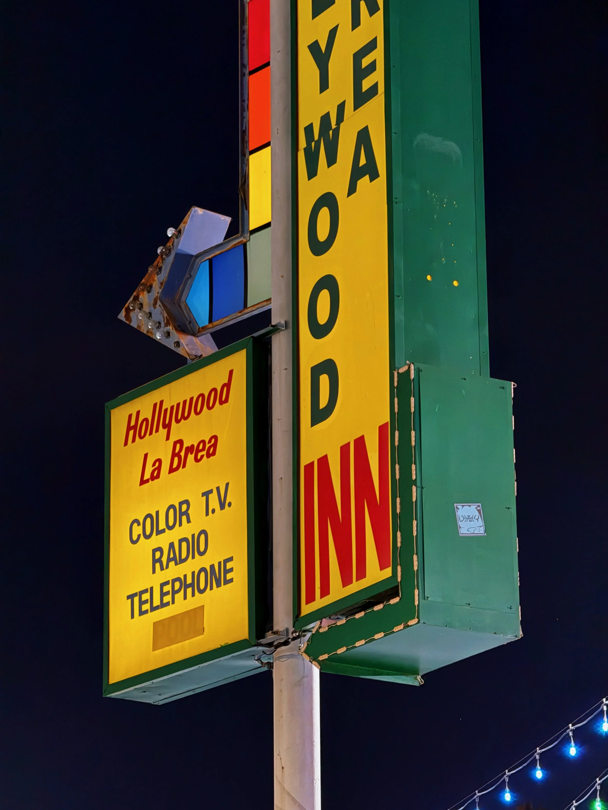 Nighttime view of a vintage-style neon-lit motel sign with yellow, red, green, and blue elements, displaying the text 'Hollywood La Brea', 'Color T.V.', 'Radio', and 'Telephone'.