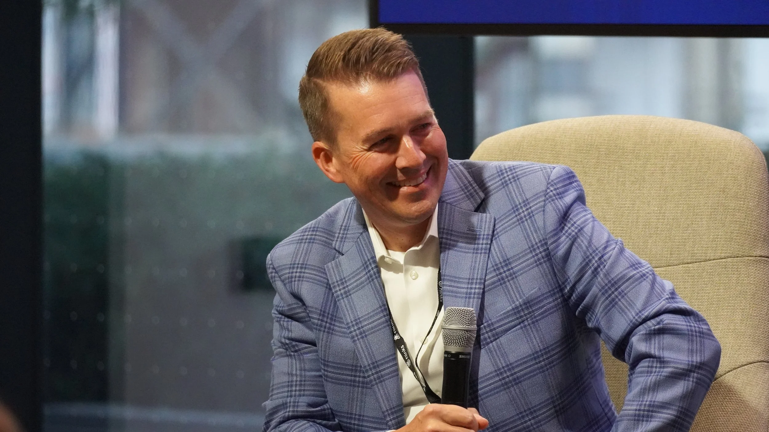 A man in a blue plaid blazer and white shirt sitting on a beige chair, smiling, holding a microphone, during a discussion or interview.