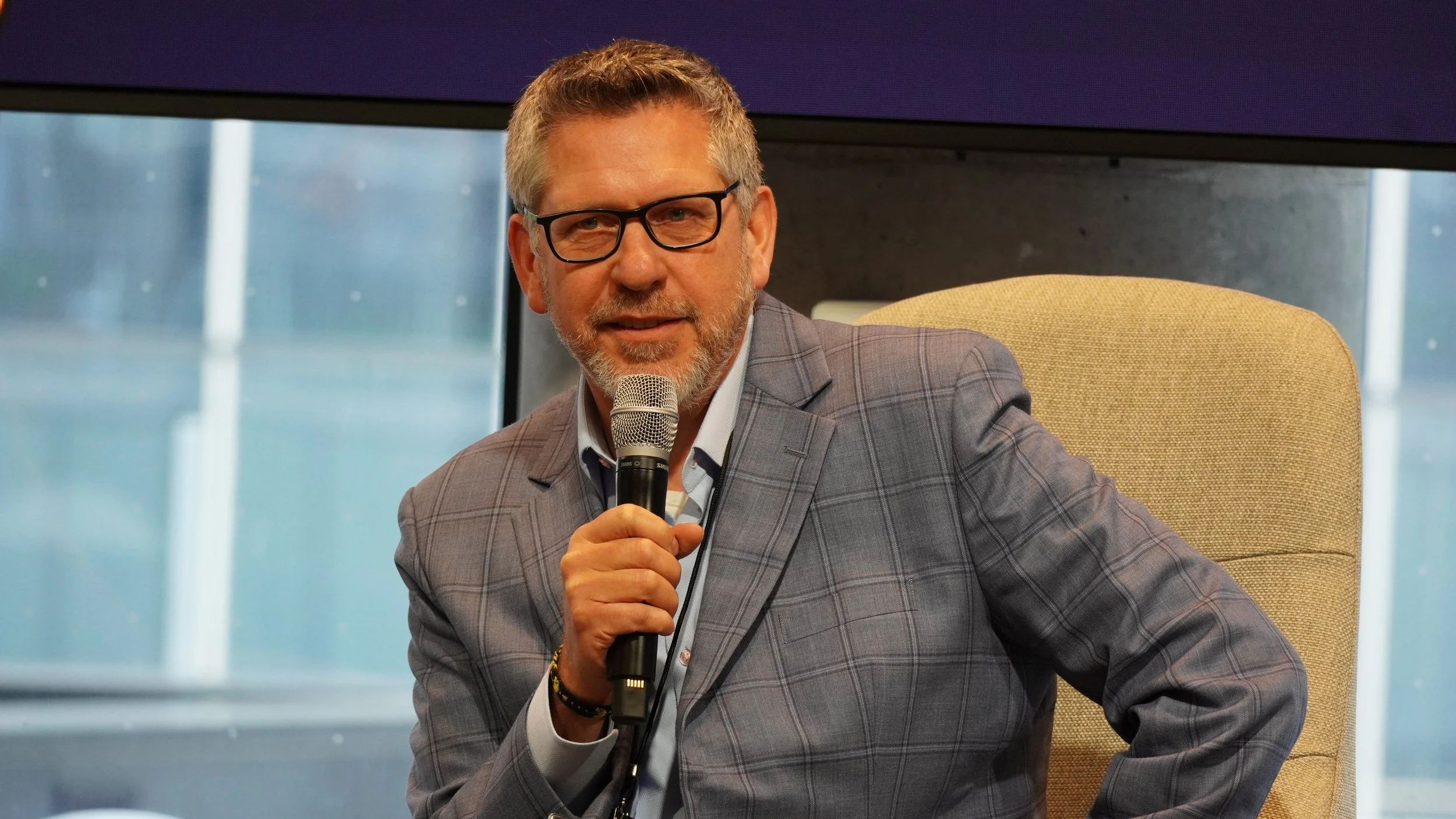 A man with gray hair and a beard wearing glasses, a gray checked suit, and a light blue shirt, sitting in a beige chair, holding a microphone, during an indoor event with large windows in the background.