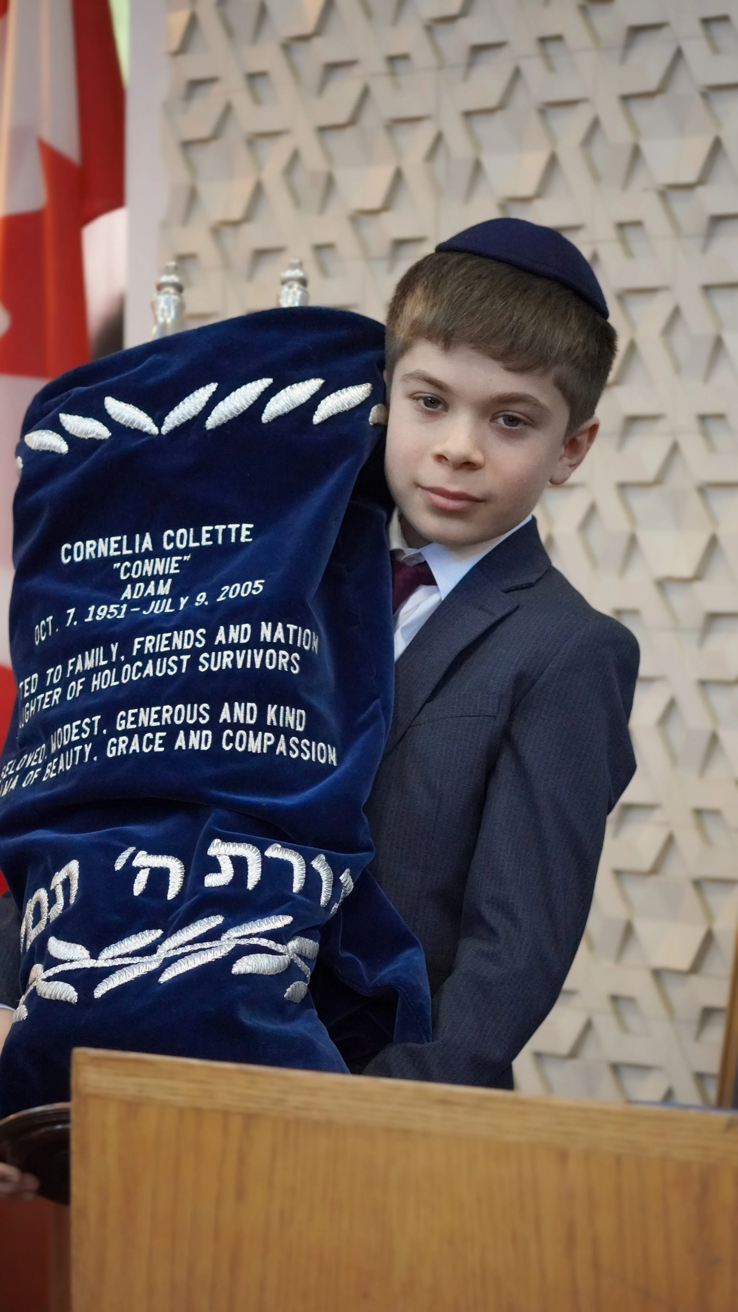 A young boy in a suit and hat holding a blue velvety memorial with white embroidered text honoring Cornelia Colette 'Connie' Adam, a Holocaust survivor, with a decorative wall behind him.