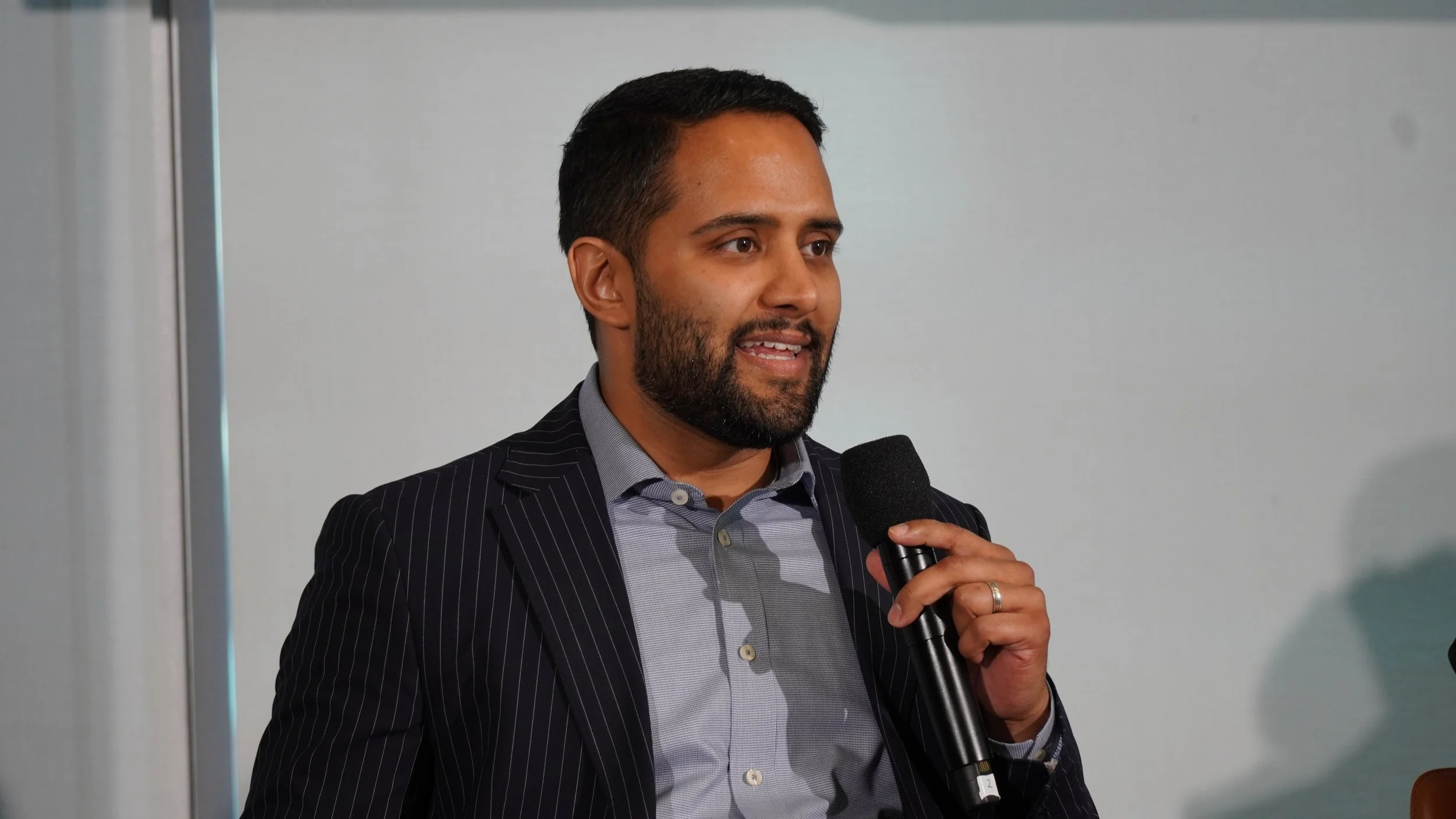 A man with a beard and short hair wearing a dark pinstripe suit and a light-colored shirt, holding a microphone and speaking at an indoor event.