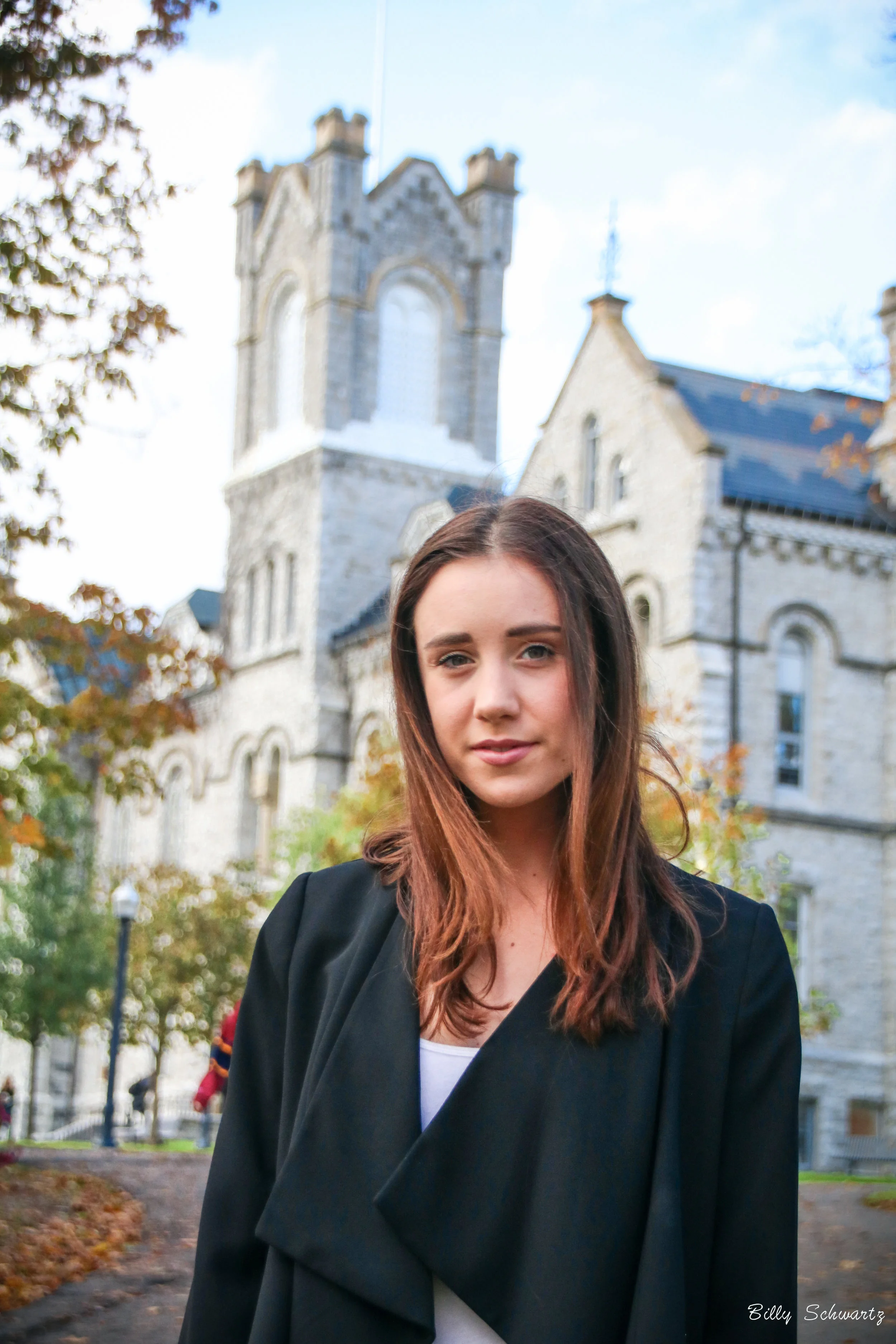 A professional corporate headshot of a young woman with brown hair standing outdoors in front of a stone building with a tall tower, surrounded by trees with autumn leaves.