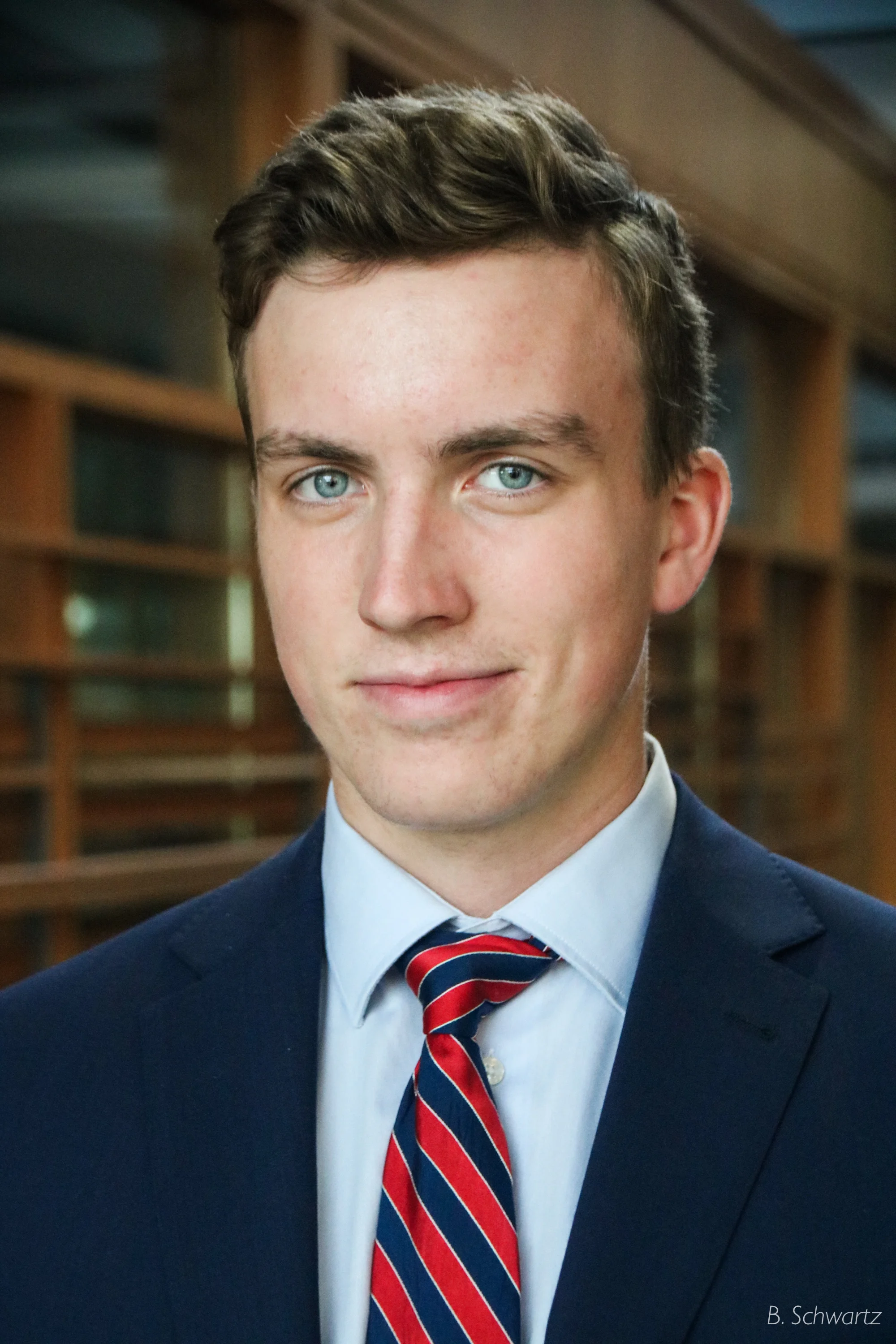 A young man in a navy blue suit, light blue shirt, and red striped tie, standing indoors with wooden shelves and books in the background.