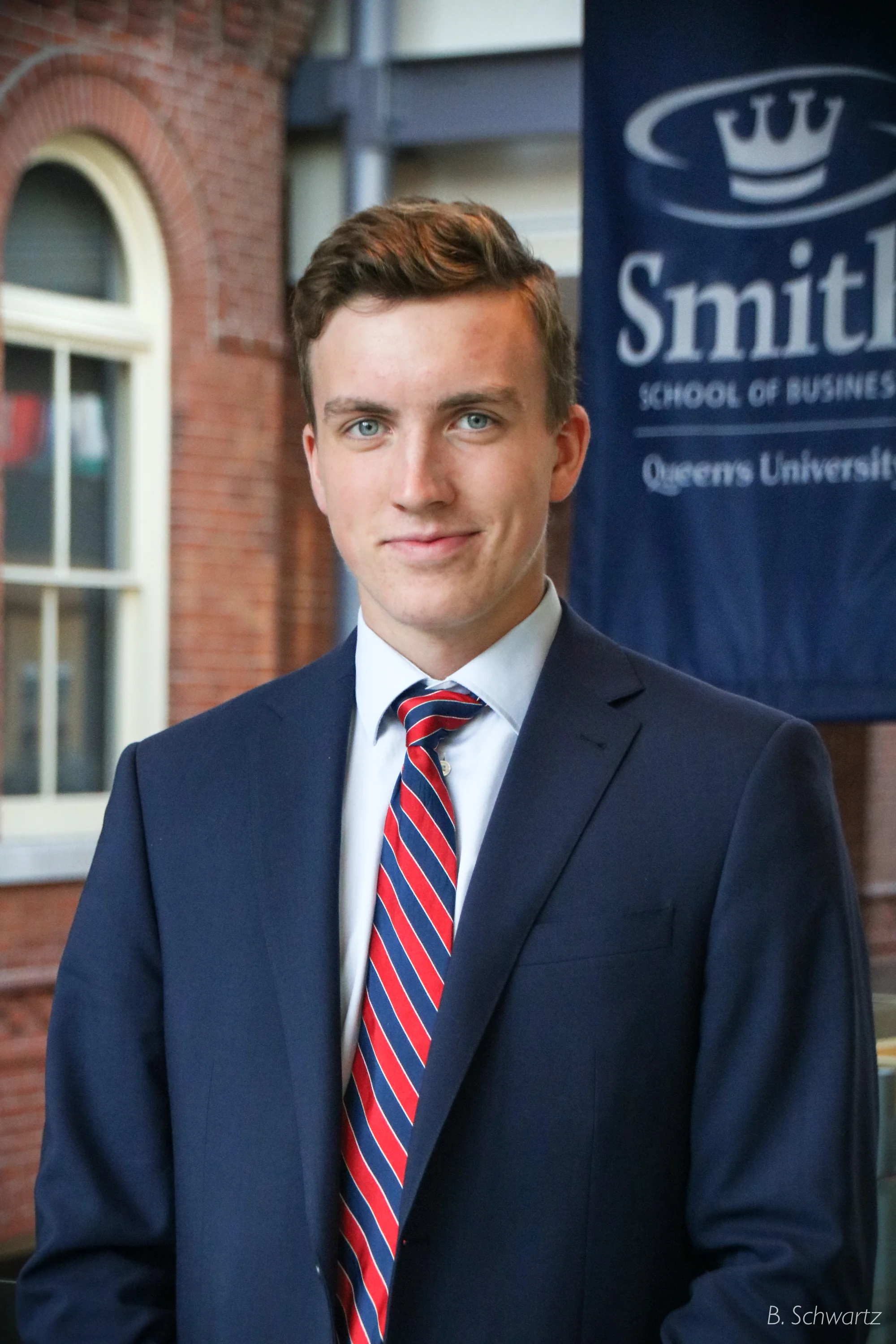 Young man in a navy suit with a white shirt and a red, blue, and white striped tie standing against a brick building with windows and a blue banner that says 'Smith School of Business, Queens University.'