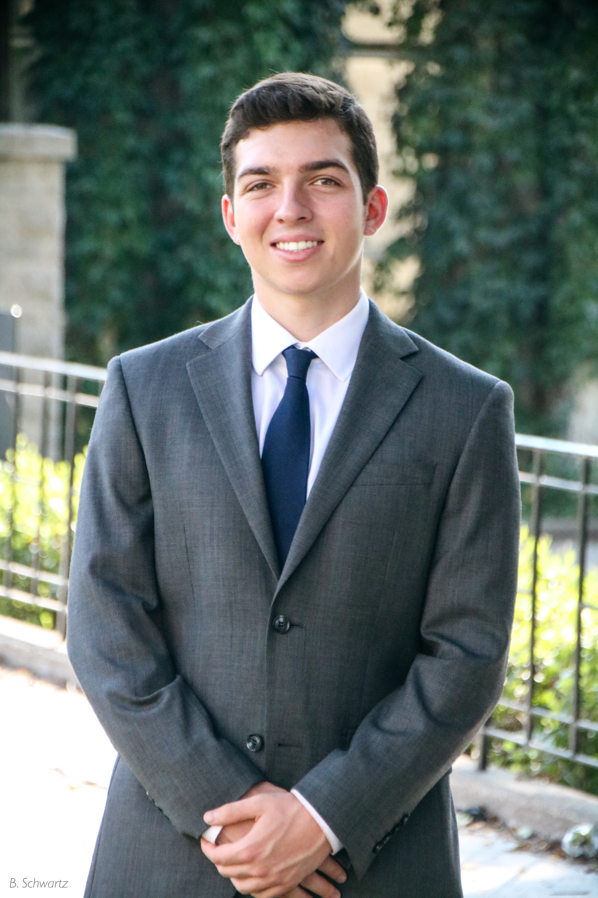 A young man in a gray suit, white shirt, and dark blue tie standing outdoors with a smile. Background includes trees and a metal fence.