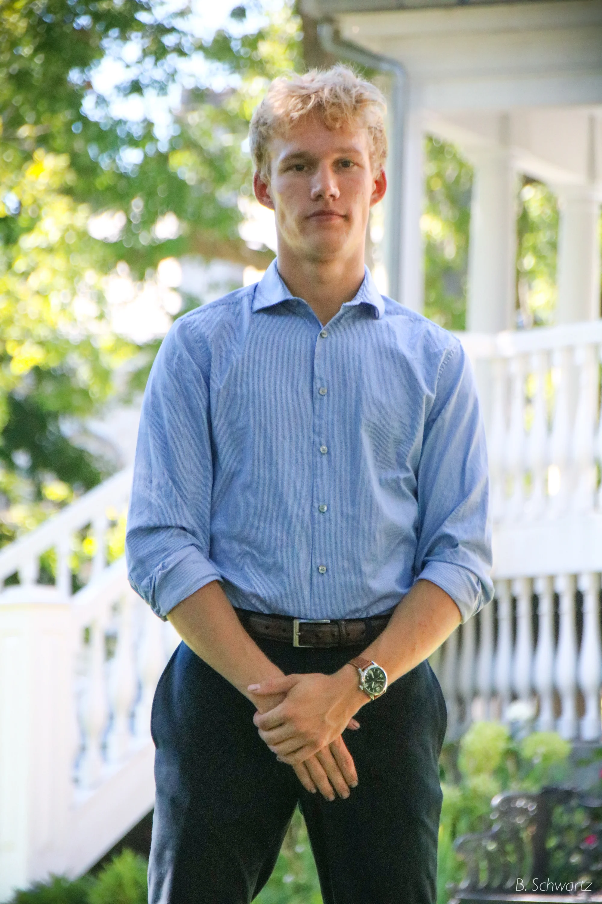 A young man with blonde hair wearing a blue shirt, black pants, and a wristwatch, standing outdoors near a white house with a porch and green trees in the background.
