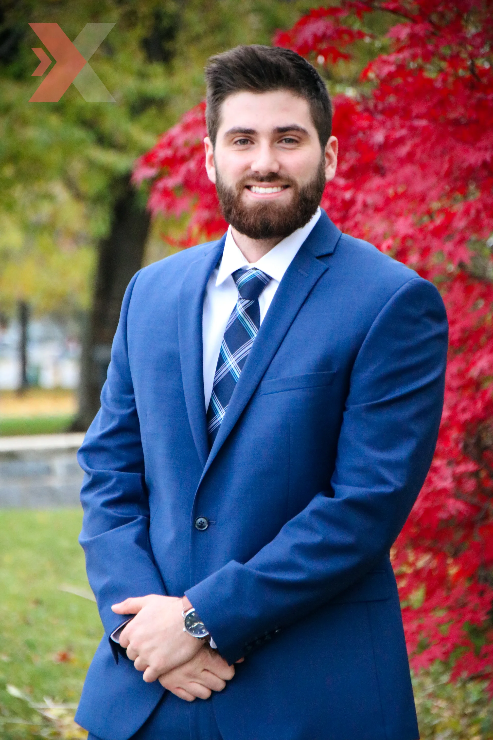 A young man with a beard in a blue suit, white shirt, and plaid tie, standing outdoors with colorful fall foliage in the background.