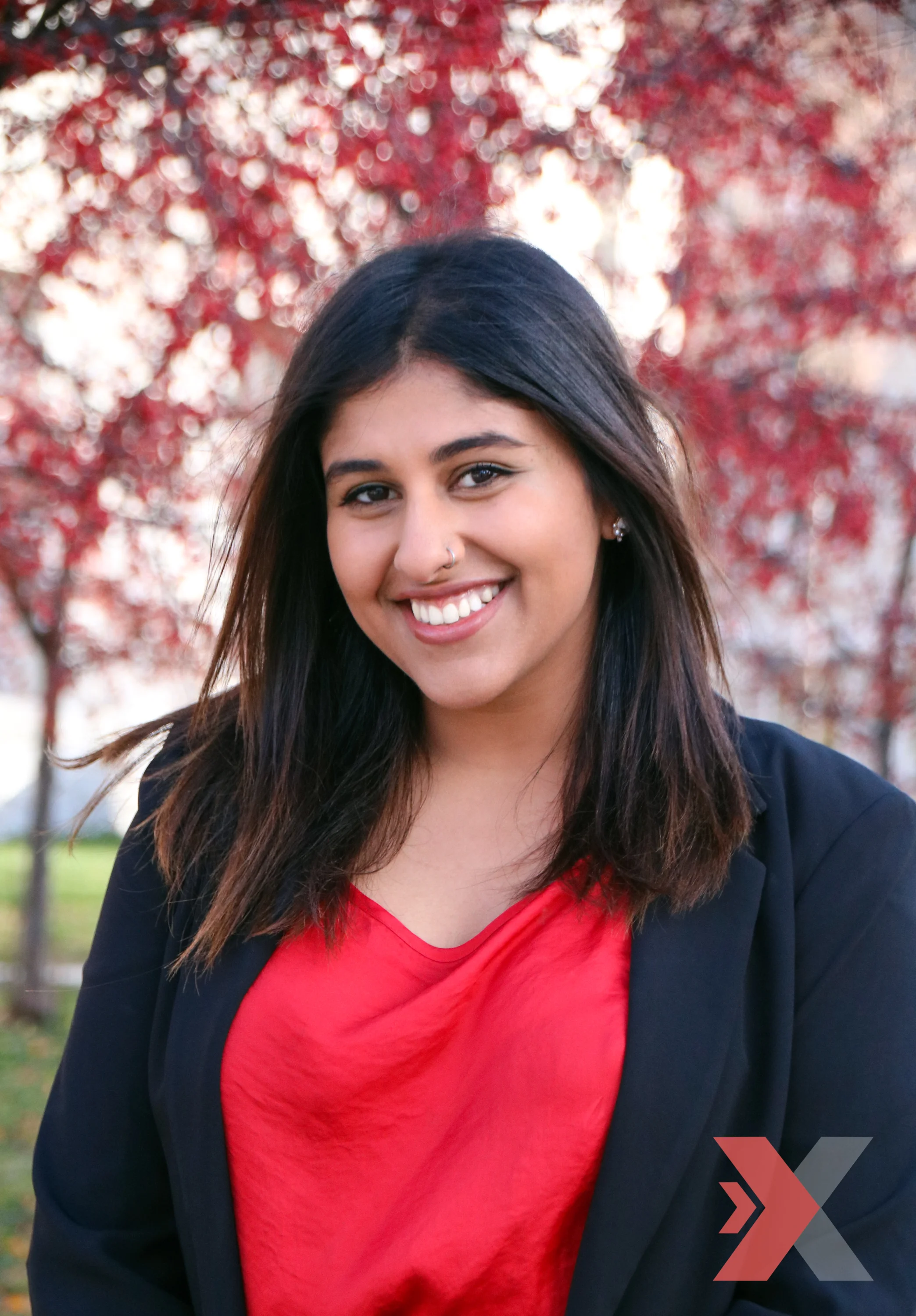 A young woman with dark hair, smiling, wearing a black blazer and a red shirt, standing outdoors with red autumn leaves in the background.