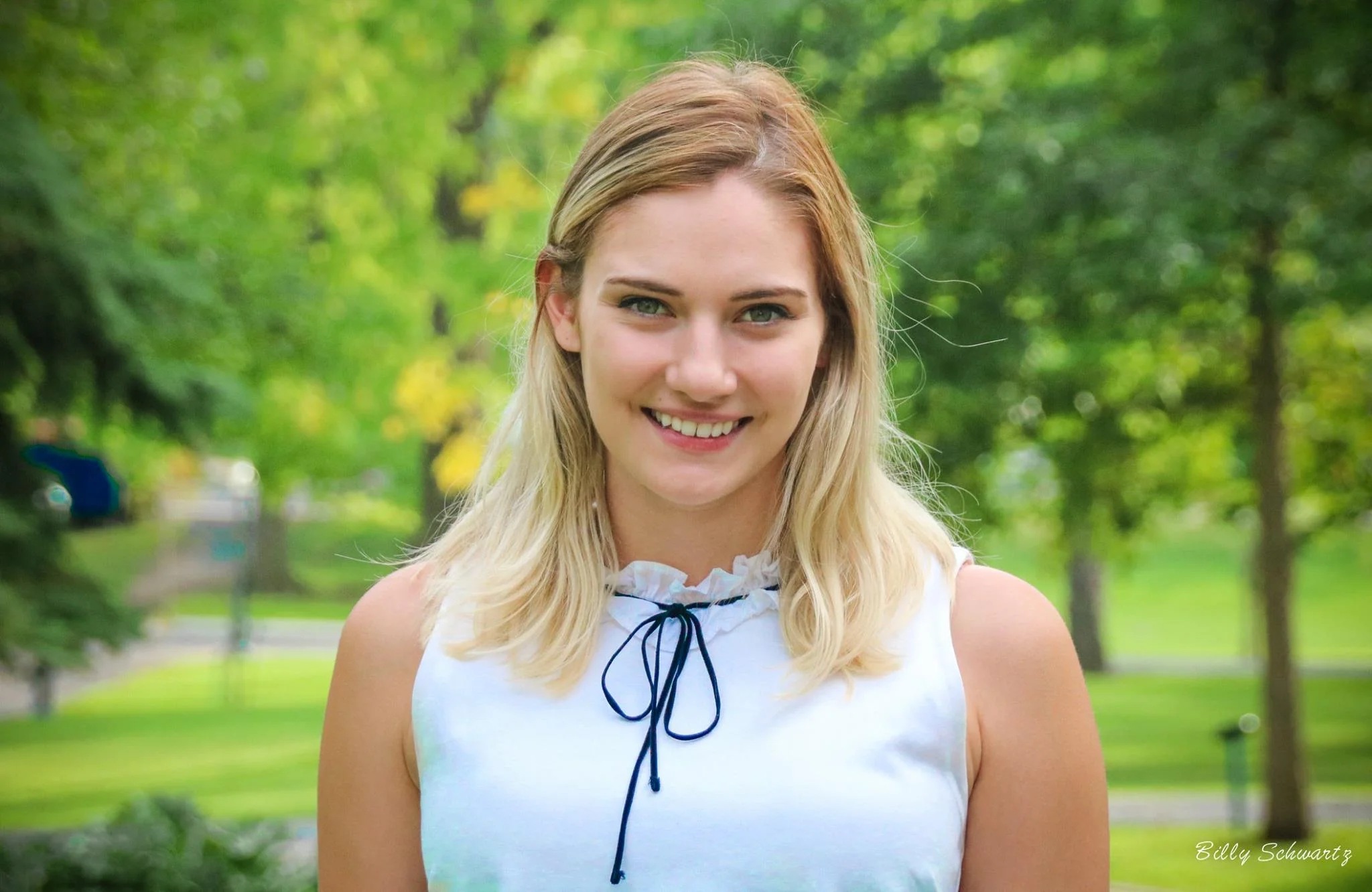 A young woman with blonde hair and blue eyes smiling outdoors in a park with green trees and grass in the background.