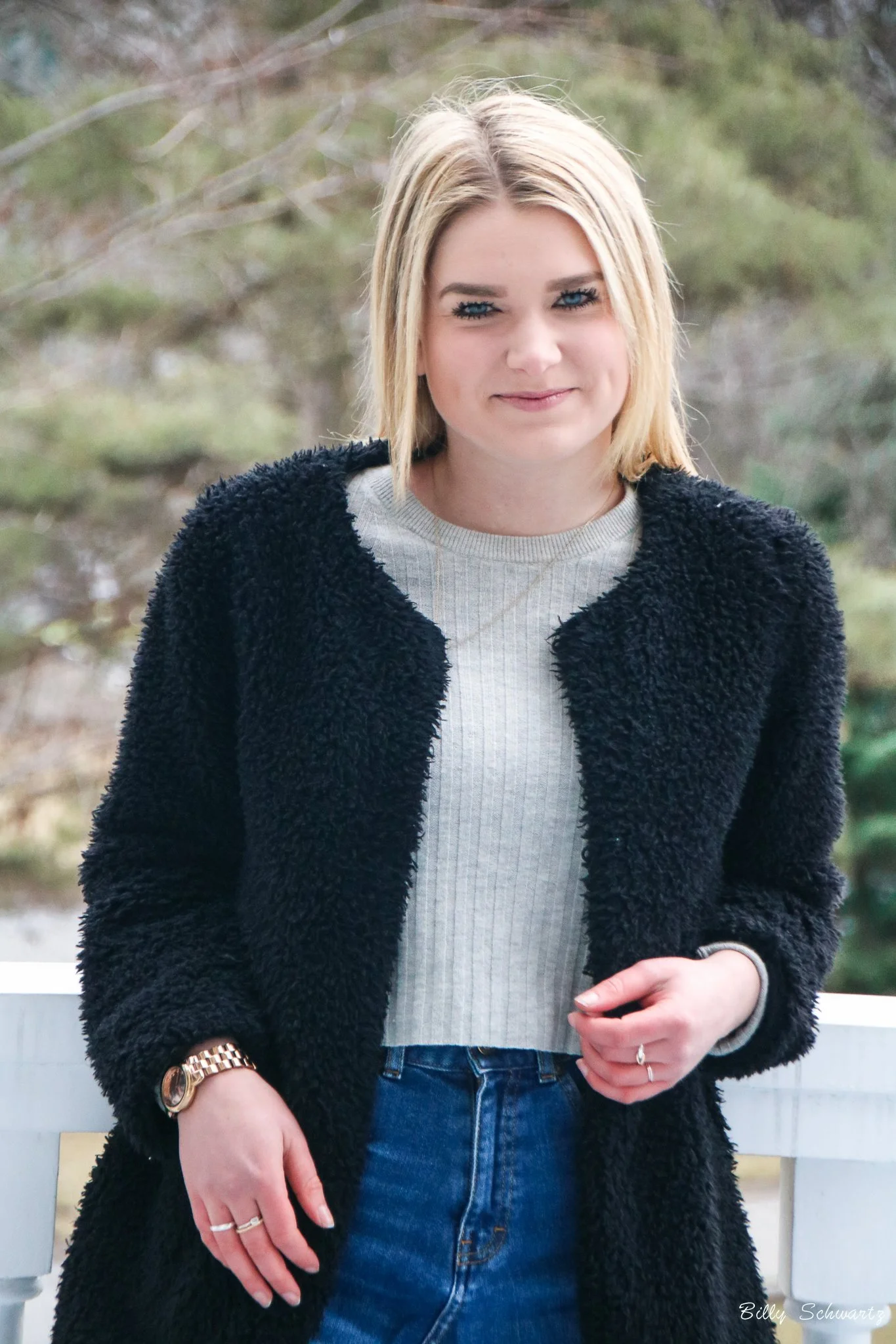 A young woman with blonde hair, blue eyes, and light skin standing outdoors with trees in the background. She is wearing a black fuzzy coat, a light gray t-shirt, and blue jeans, with a watch and rings on her left hand.