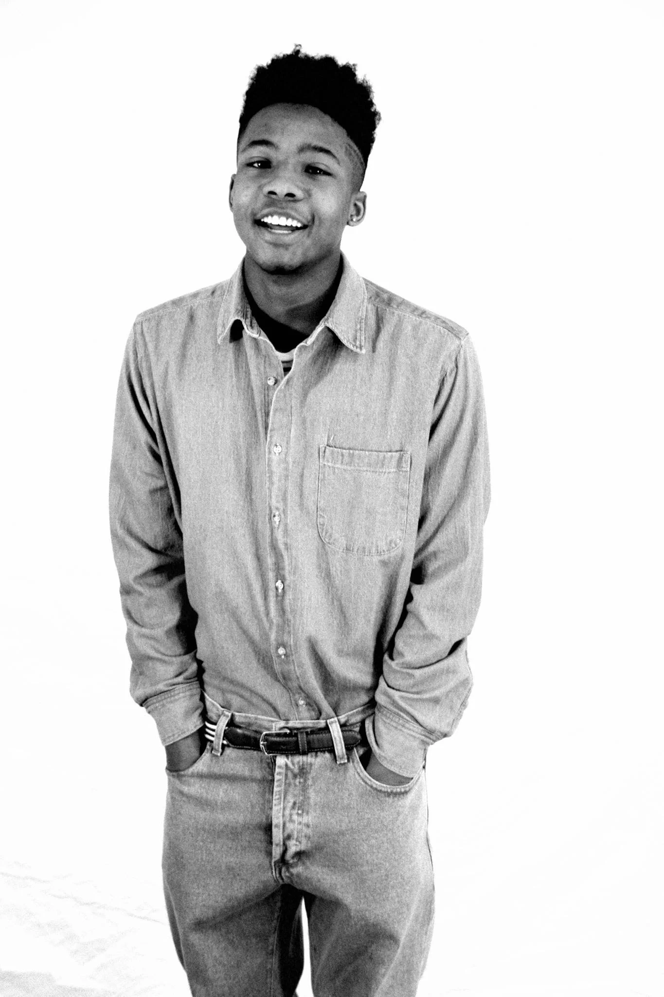 Black and white fashion photo of a young man smiling, wearing a denim shirt and jeans with hands in his pockets, standing against a plain background.