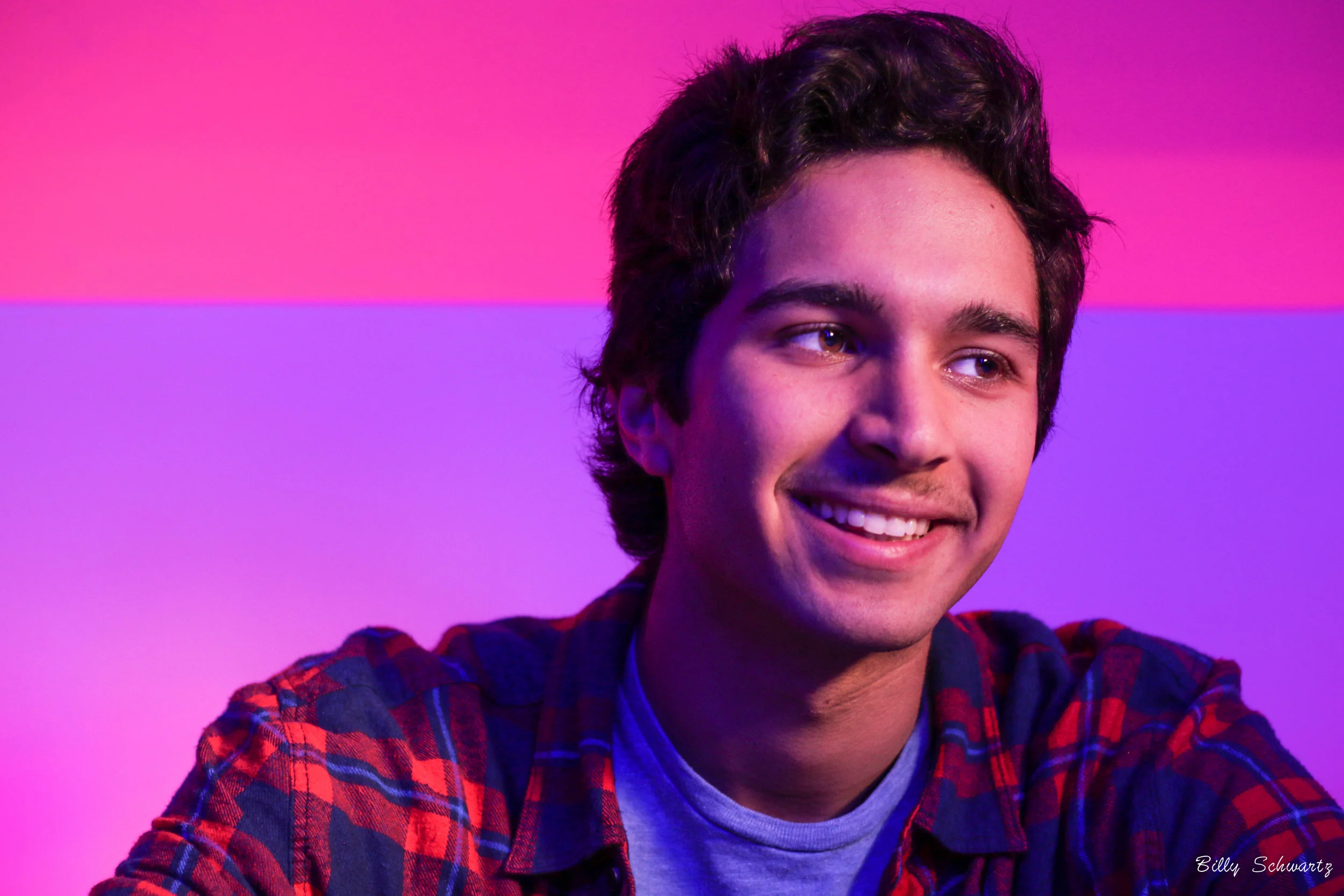 Young man with dark hair smiling, wearing a checkered shirt, against a vibrant pink and purple background.