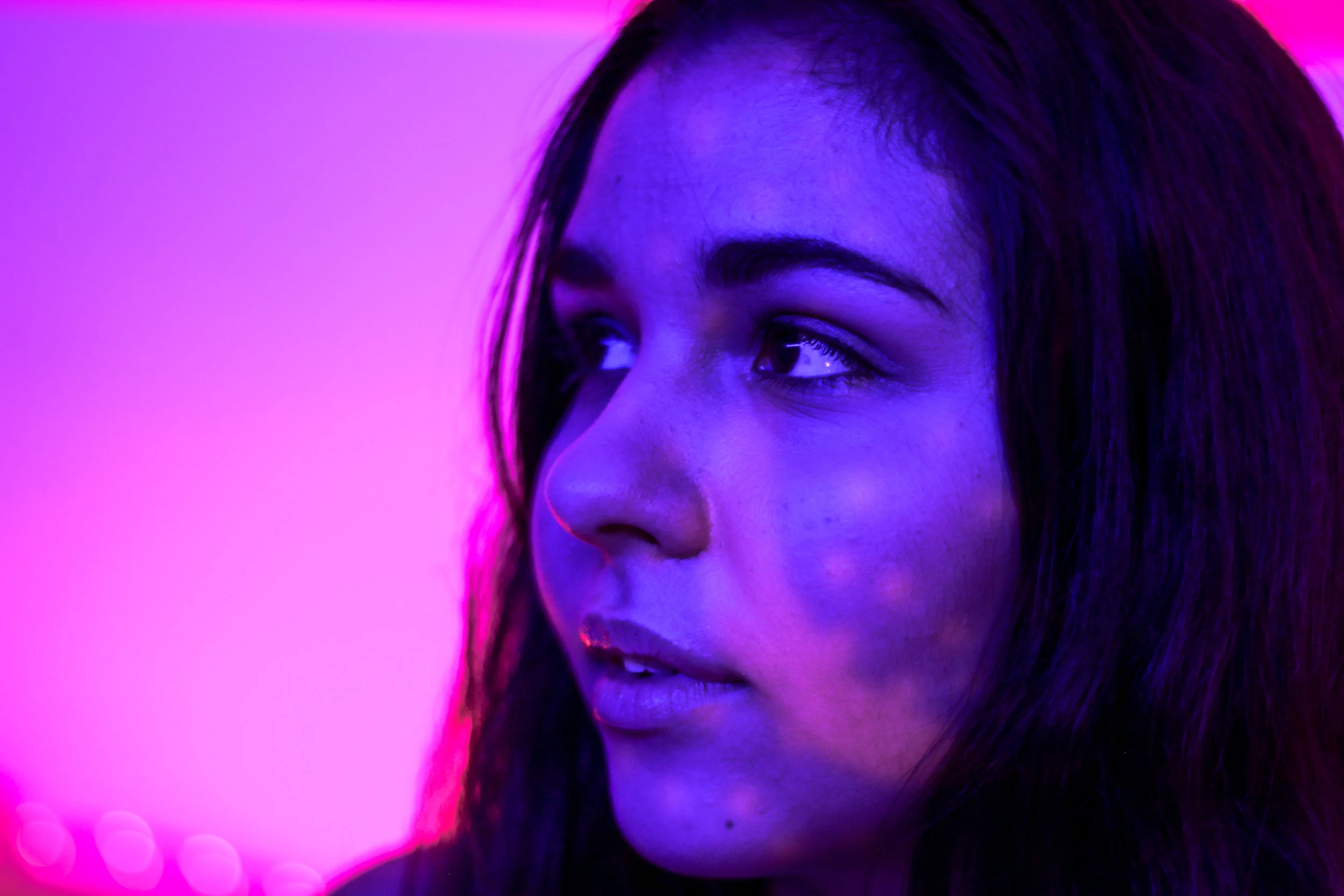 A woman with dark hair looking to the side, illuminated by blue and pink neon lights.