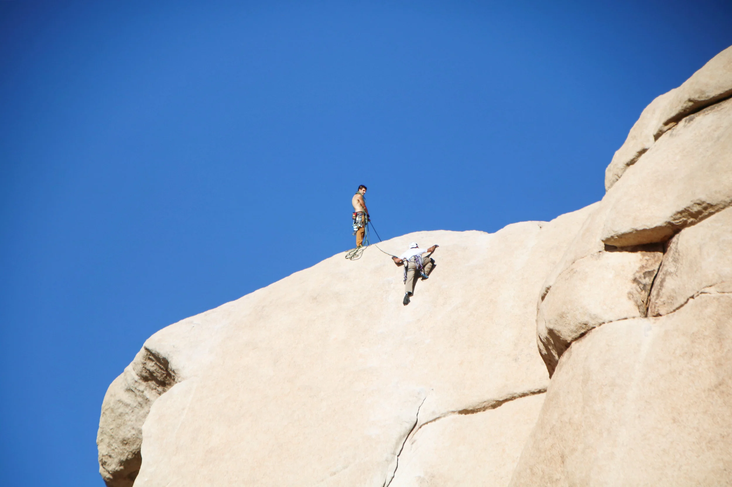 Two rock climbers on a large granite boulder with a clear blue sky in the background. One climber is hanging from the rock, while the other is standing and holding the climbing rope.