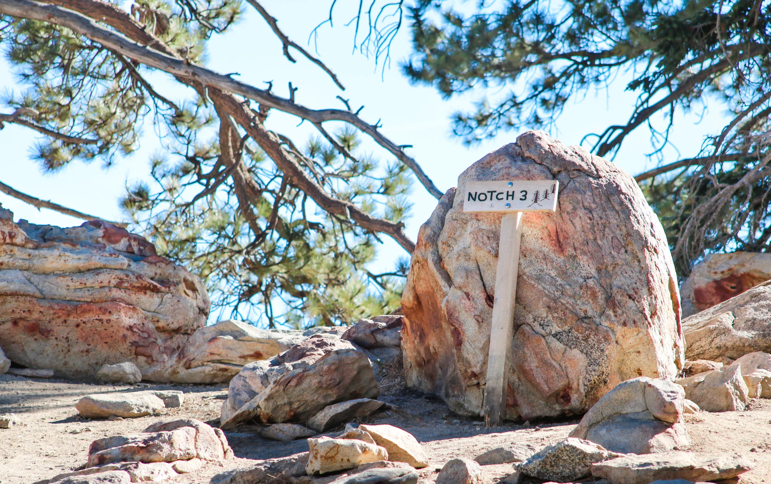 Large rock in a natural outdoor setting with a sign that reads 'NOTCH 3' and depicts two trees, with other rocks and pine trees in the background under a clear blue sky.