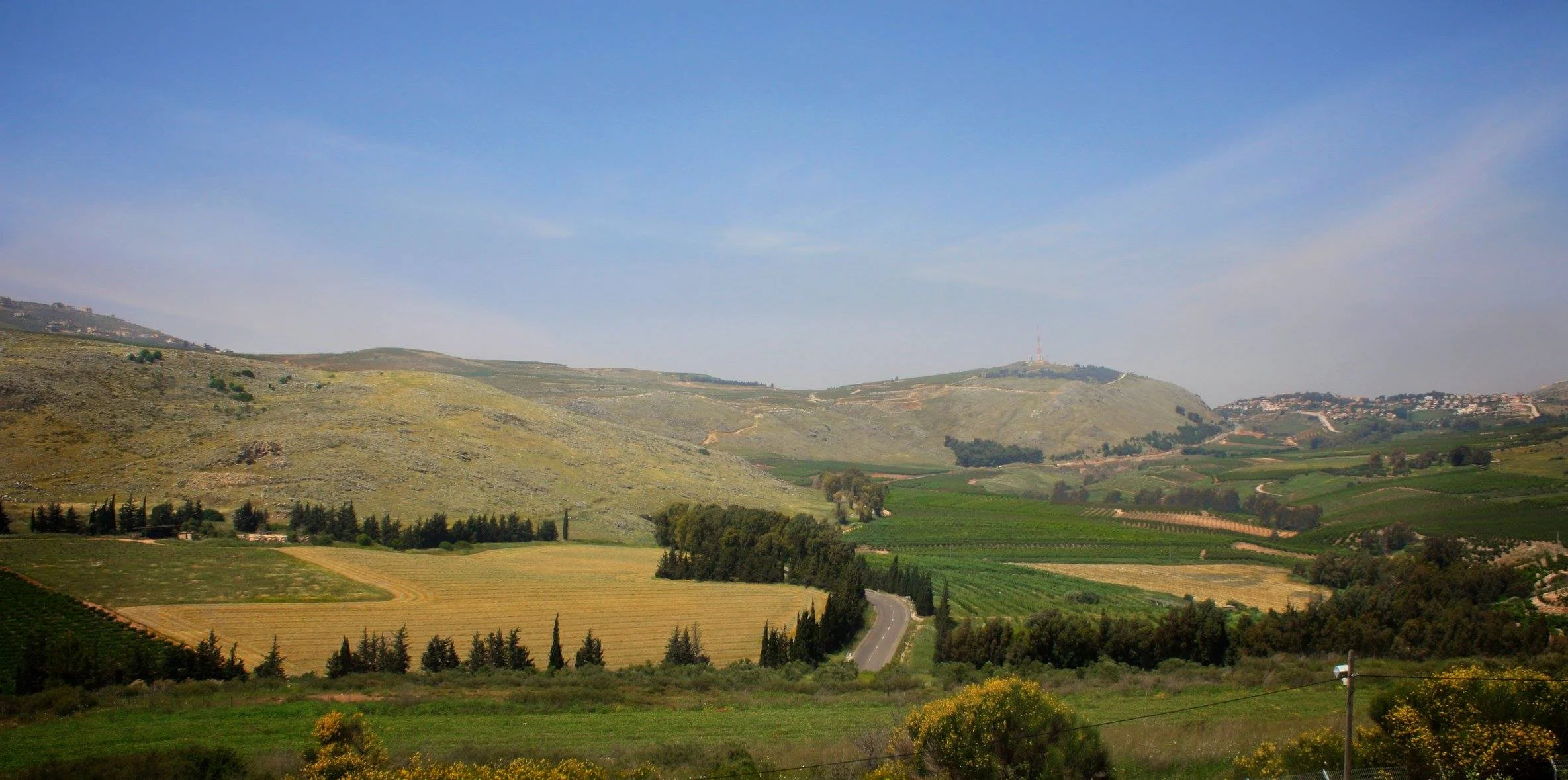 Scenic view of rolling green hills, farmland, and a winding road under a blue sky.