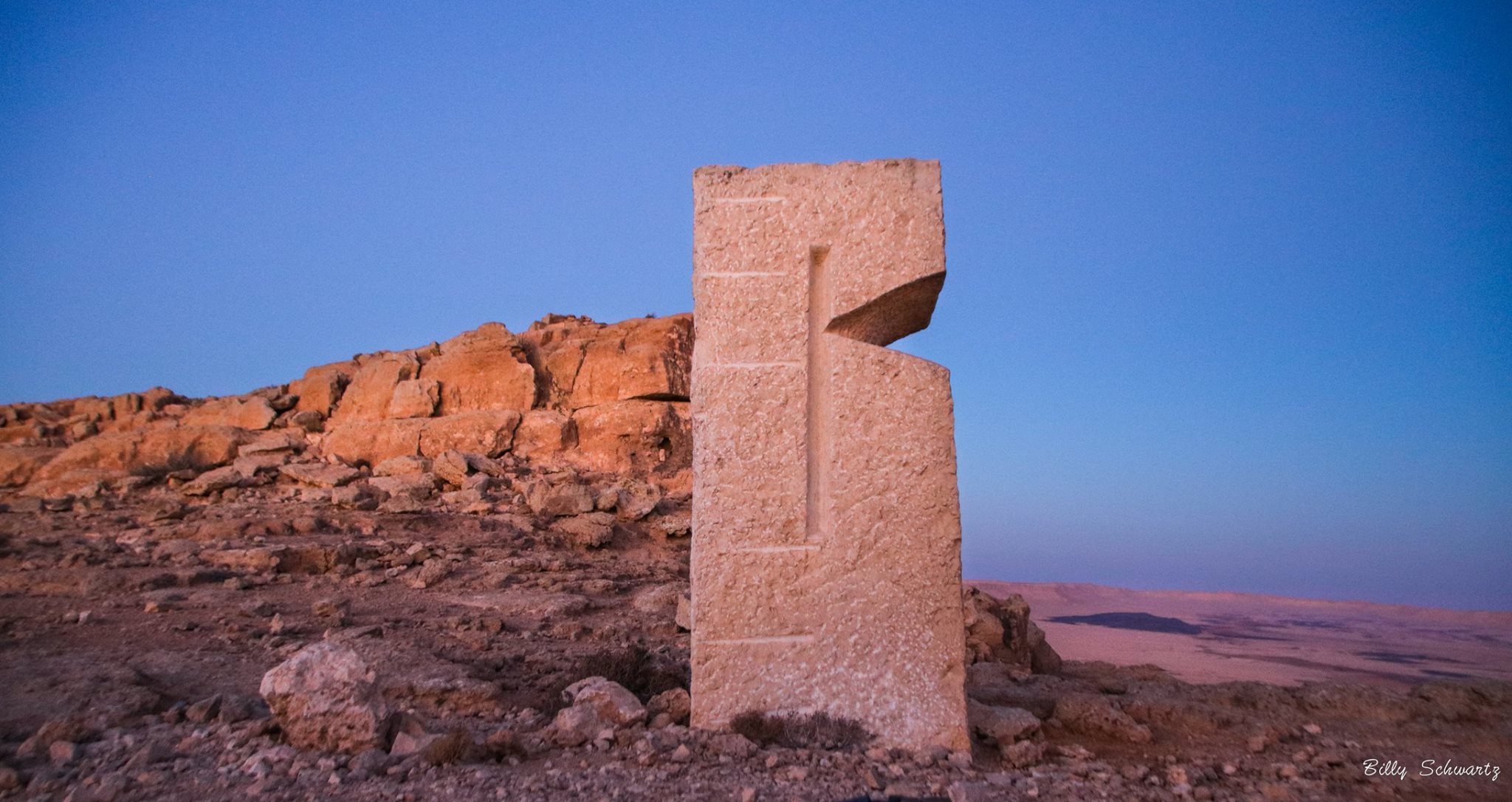 A tall, rectangular stone monument with a cutout in the shape of the letter 'F' stands on rocky terrain against a clear blue sky at sunset.