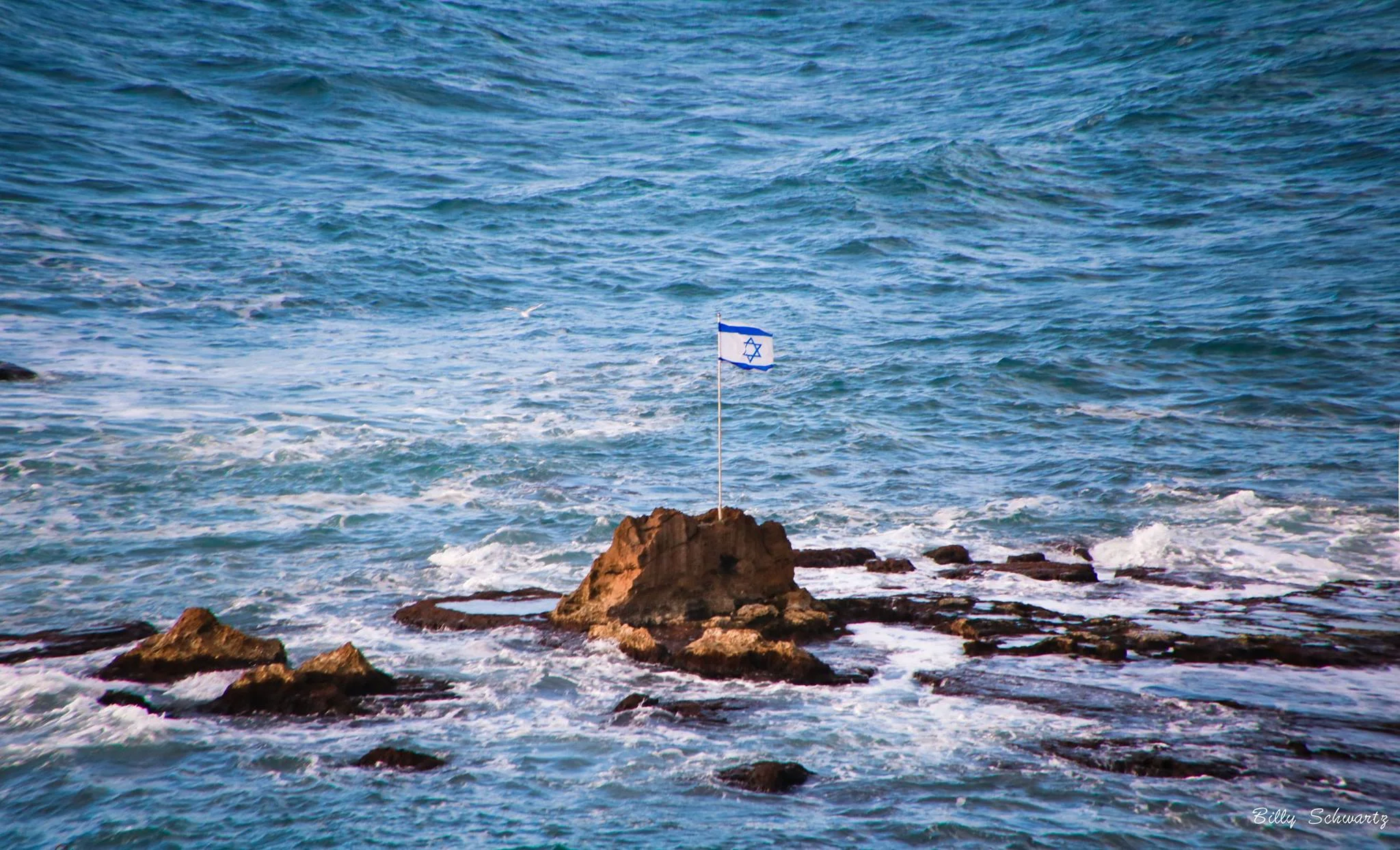 An ocean view with a rock formation in the water displaying a small Israeli flag on a pole.