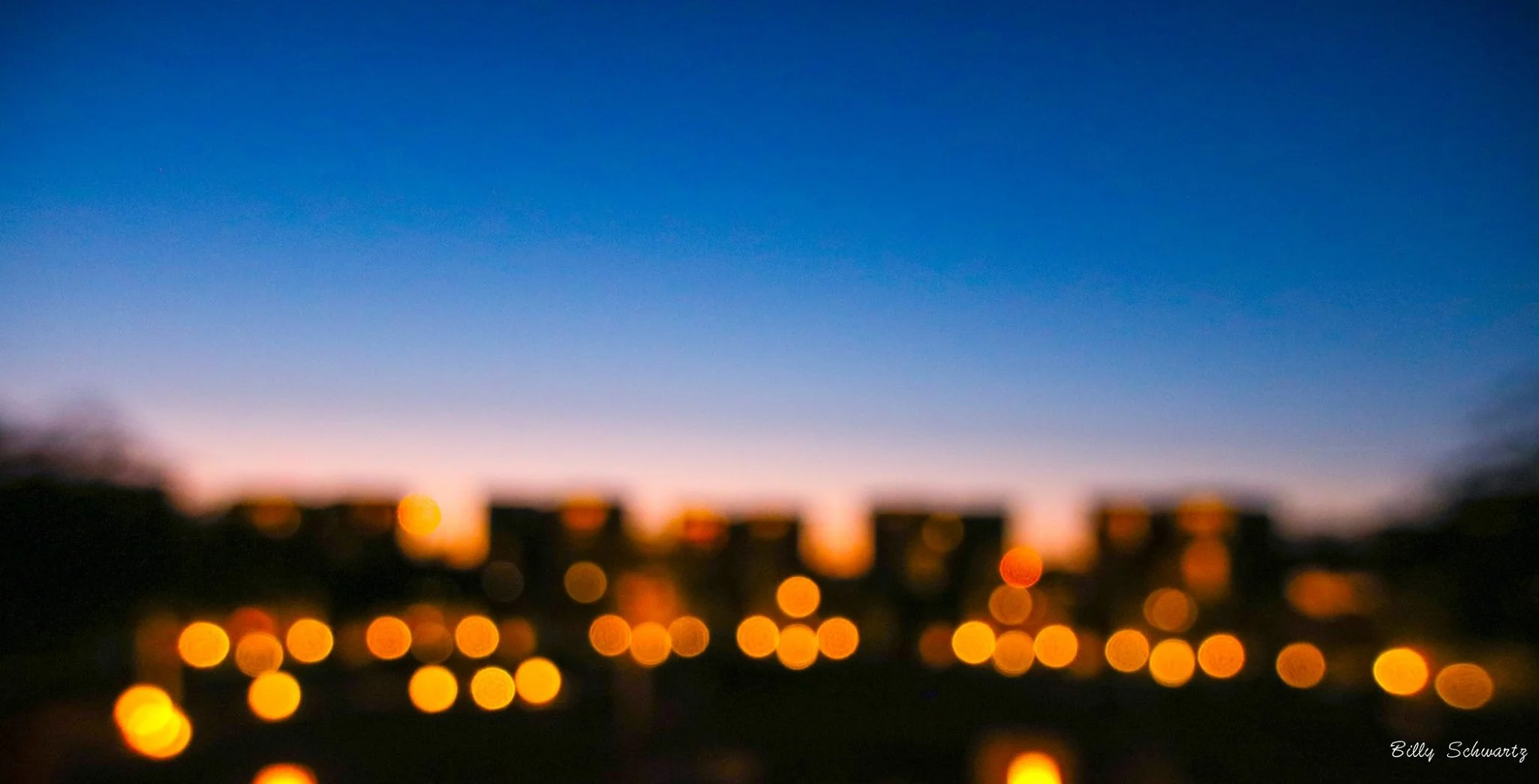 Blurry photograph of a cityscape at dusk with a colorful sky transitioning from orange to deep blue, and out-of-focus city lights creating bokeh effects.