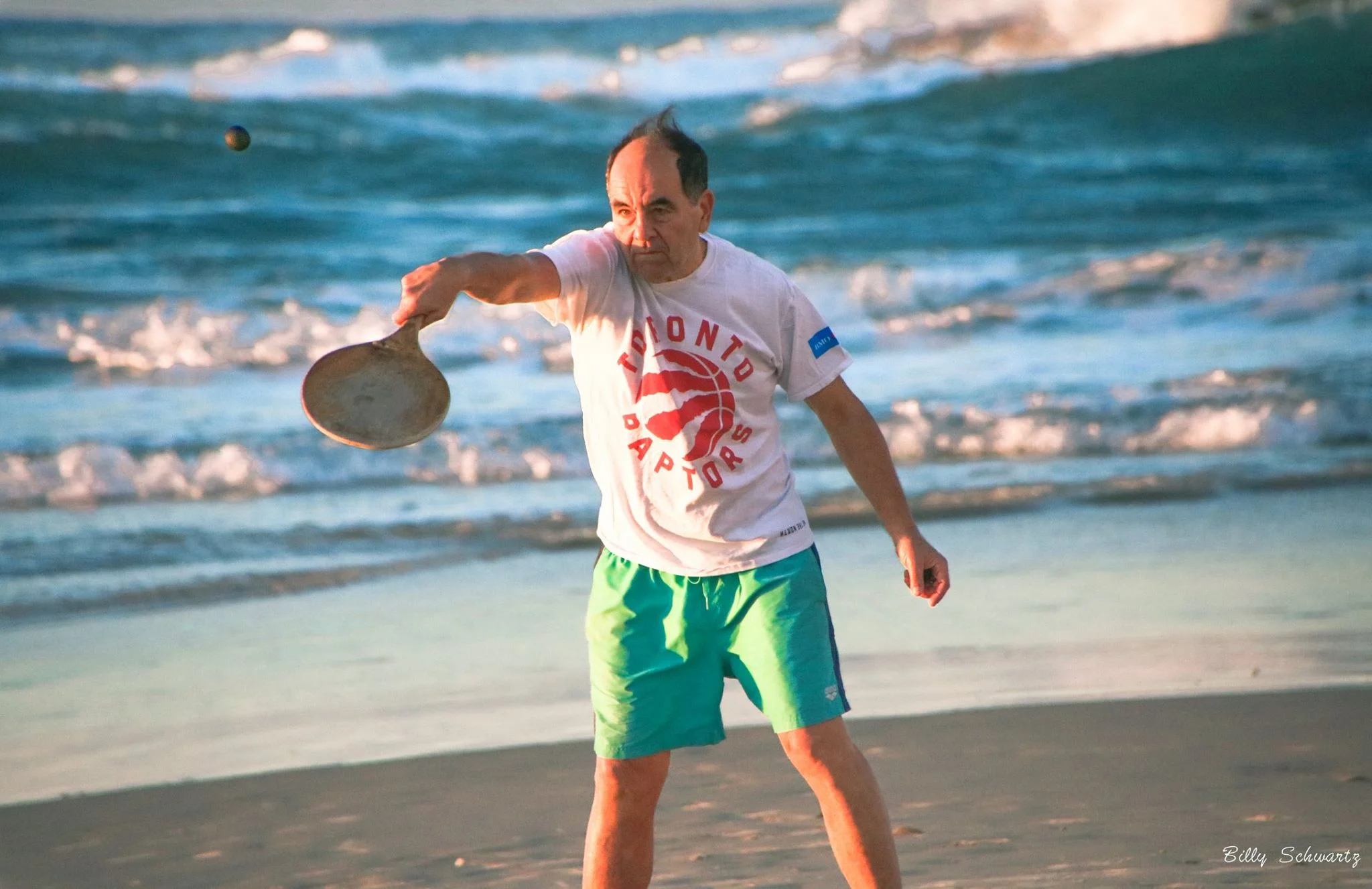 A man playing paddleball on a beach, wearing a Canada Toronto Raptors t-shirt and green shorts, with ocean waves in the background.
