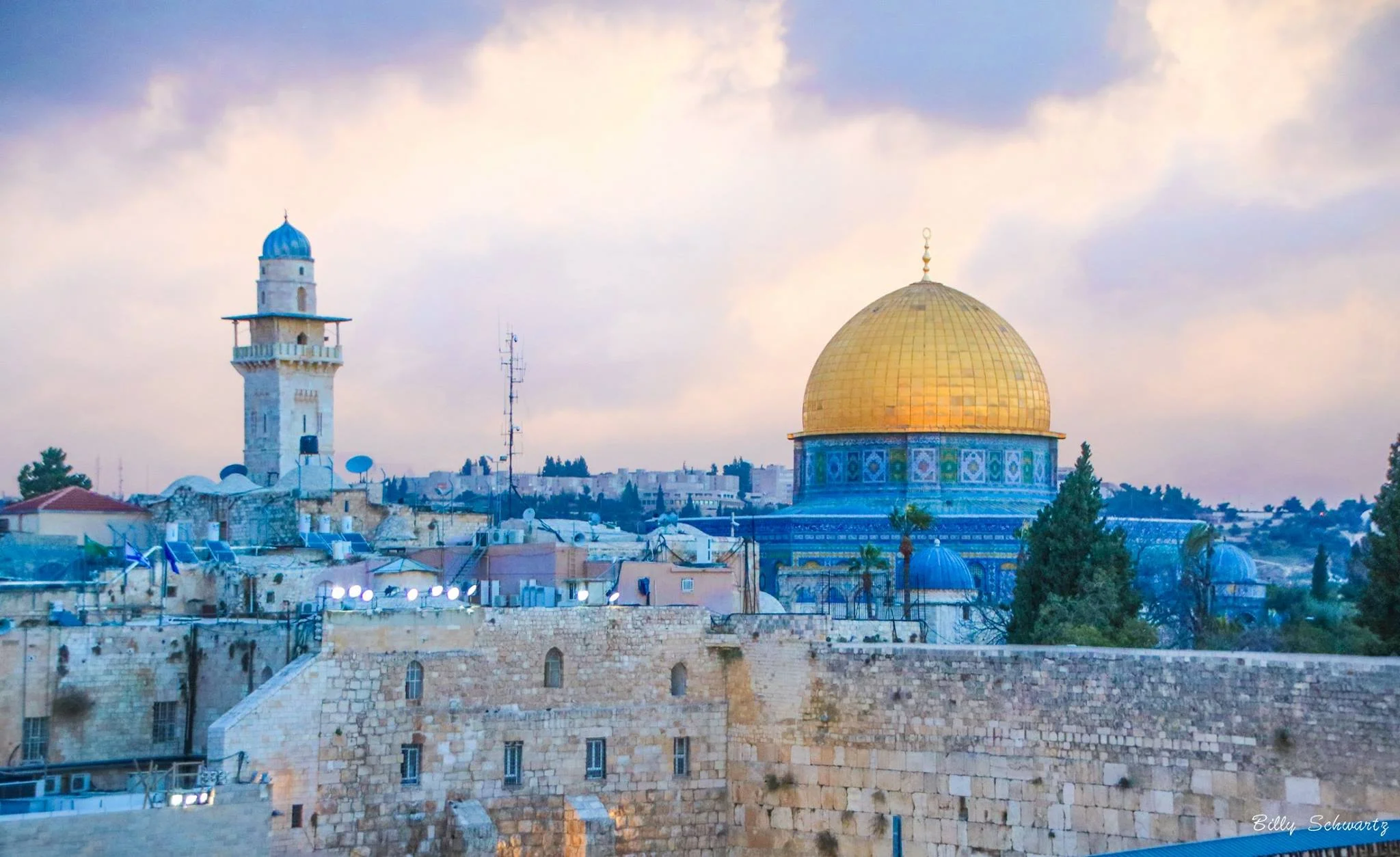 View of the city of Jerusalem featuring the Dome of the Rock with a shining golden dome, surrounded by historic stone buildings and a cloudy sky.