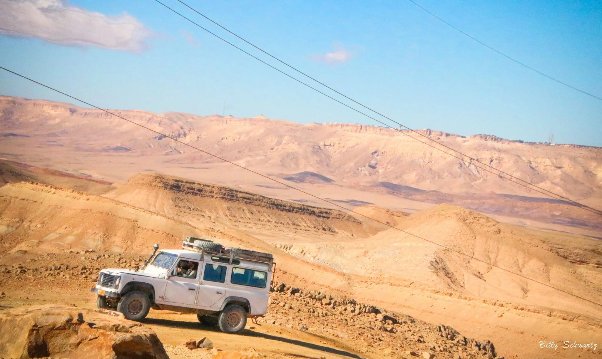A white off-road vehicle on a dirt road in a desert landscape with mountains in the background under a blue sky.