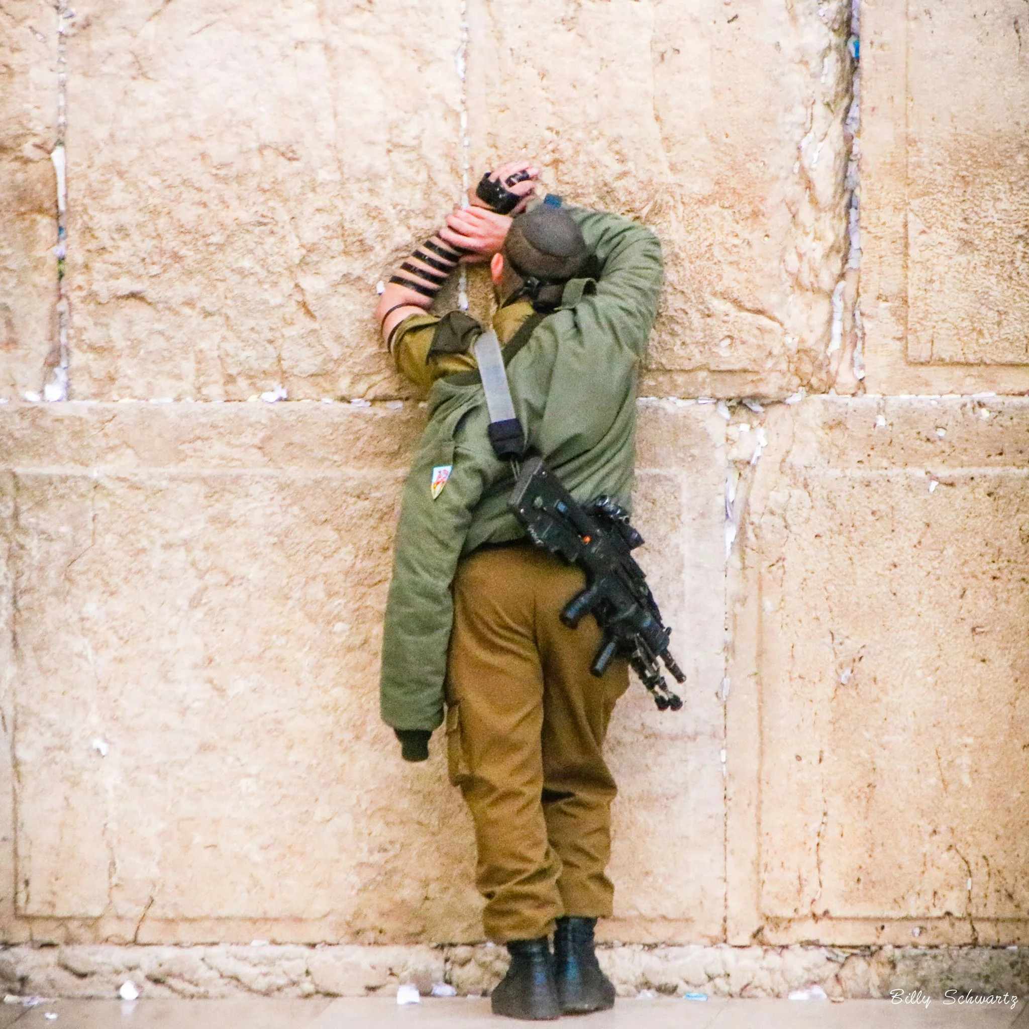 Person leaning against a stone wall with hands pressed to the wall, wearing military-style clothing and gear, including a rifle strapped to their side.