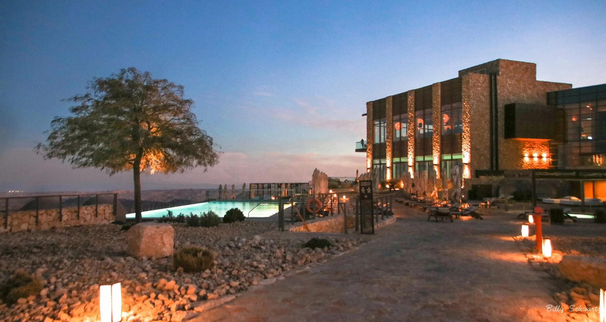 An outdoor scene at dusk featuring a modern stone and glass building with illuminated windows, a tree, a small pool with underwater lights, several lounge chairs, and pathway lights.