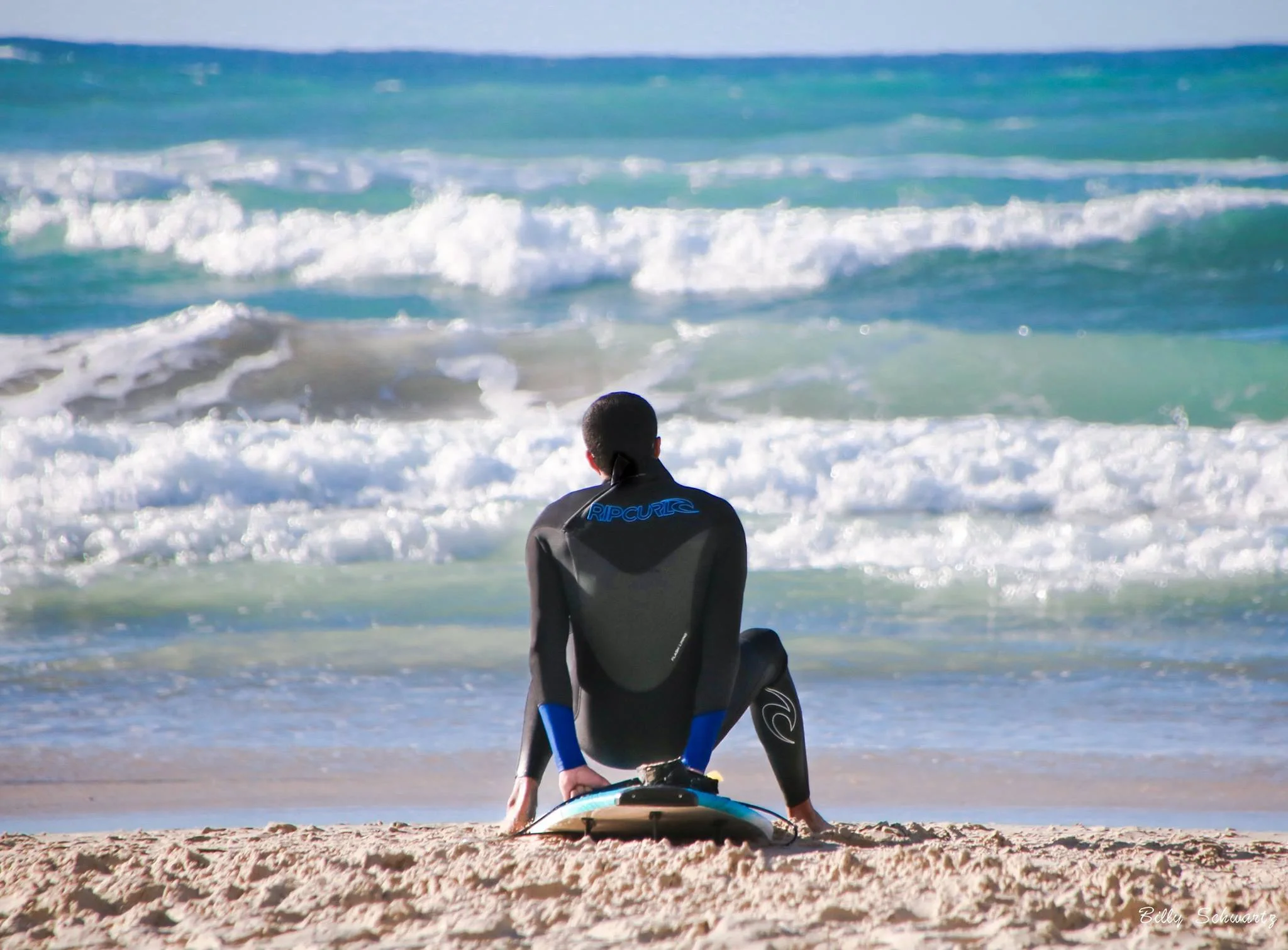 A person in a wetsuit sitting on the sandy beach, facing the ocean waves, preparing for surfing.
