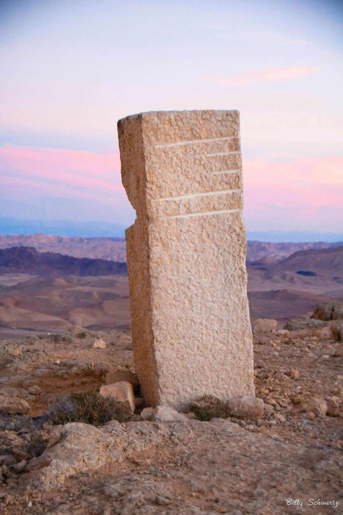 A large stone standing upright outdoors with a mountainous landscape and pastel sky in the background.