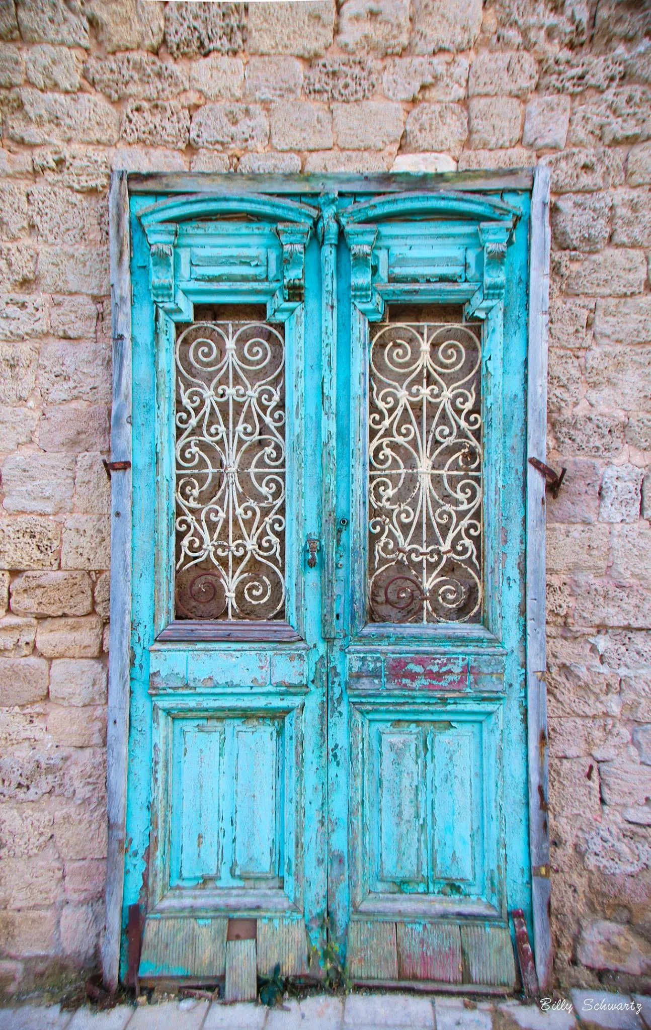 An old, weathered turquoise wooden door with ornate white wrought iron grilles in a stone wall.