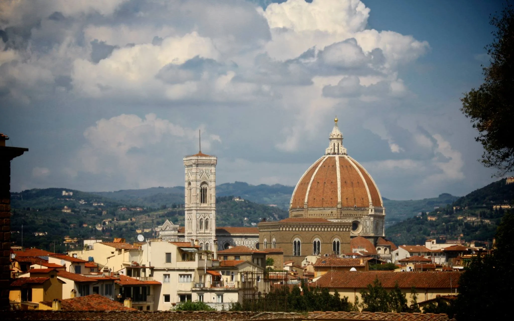 View of Florence, Italy, featuring the iconic Florence Cathedral with its large red-tiled dome and the bell tower, set against a backdrop of rolling hills and a cloudy sky.