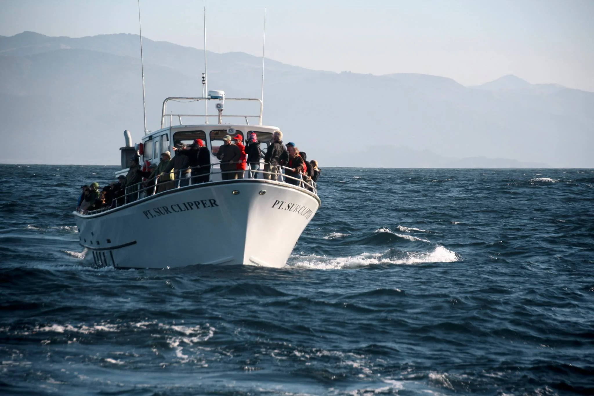 A boat named PT SUR CLIPPER with multiple passengers on board, moving through the water with distant mountains in the background.