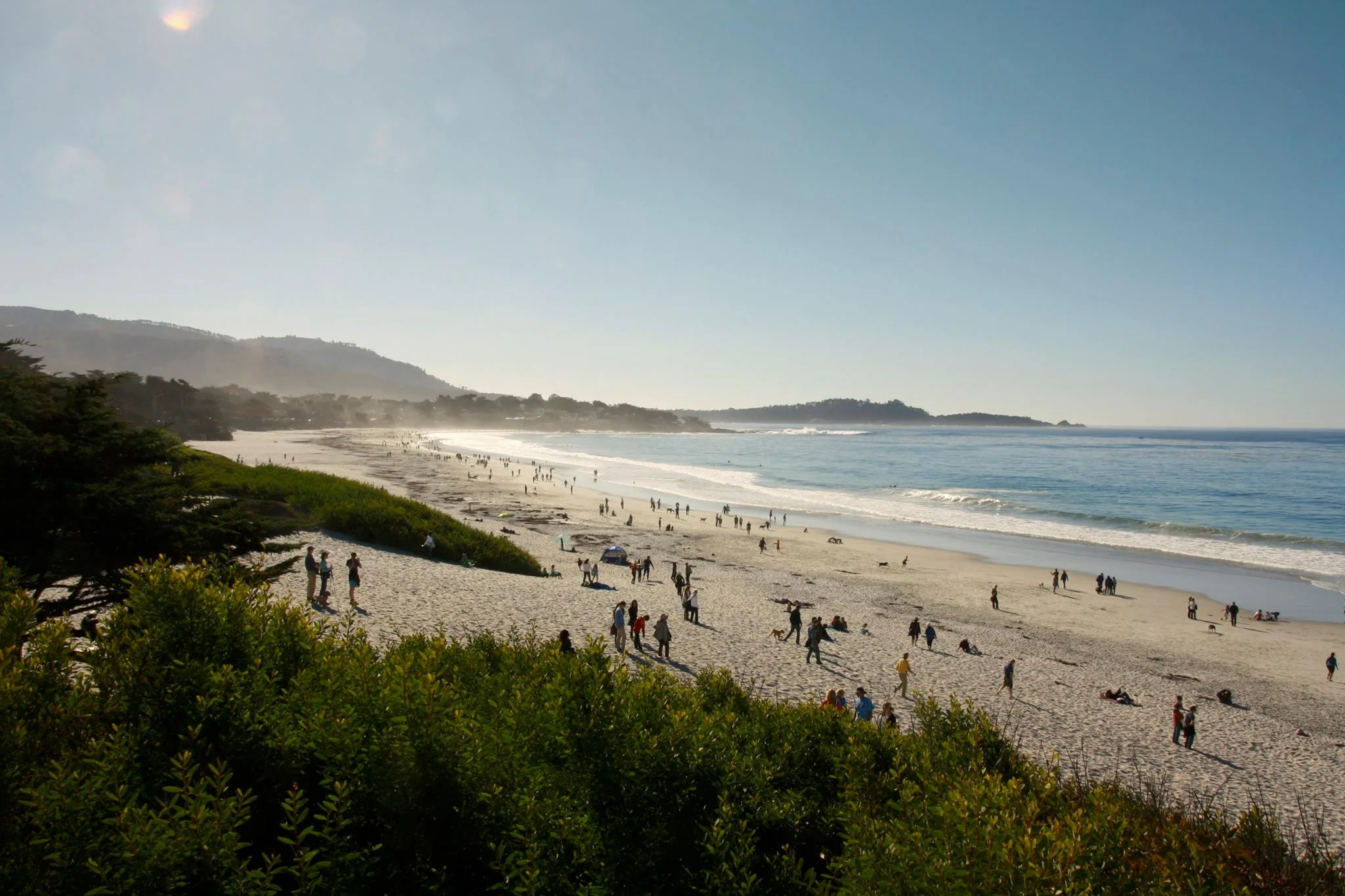 A crowded beach with many people walking and sitting on the sand, ocean waves, and a distant headland under a clear blue sky with some fog near the horizon.