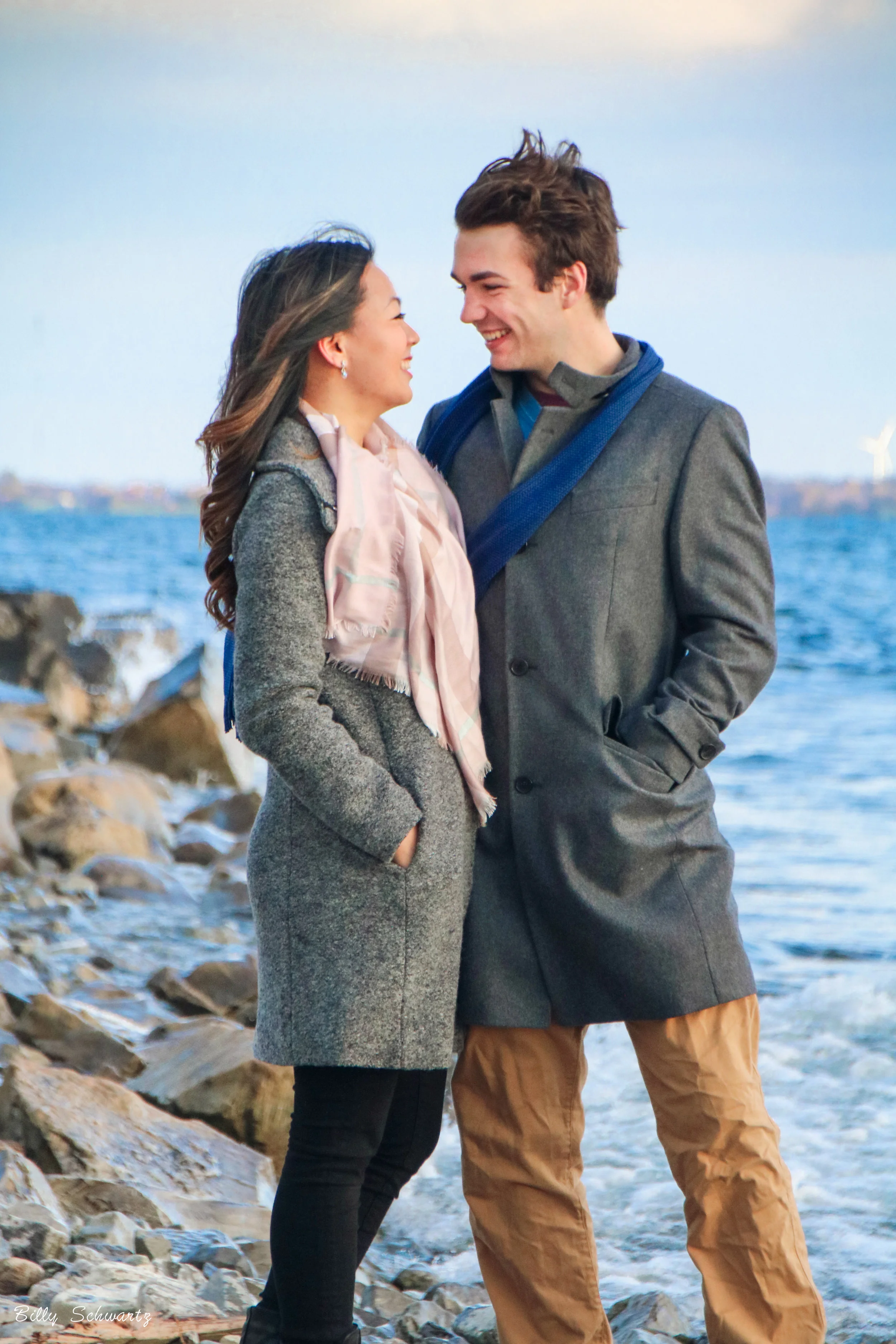 A young couple smiling and looking at each other near a rocky shoreline with the ocean and a wind turbine in the background.