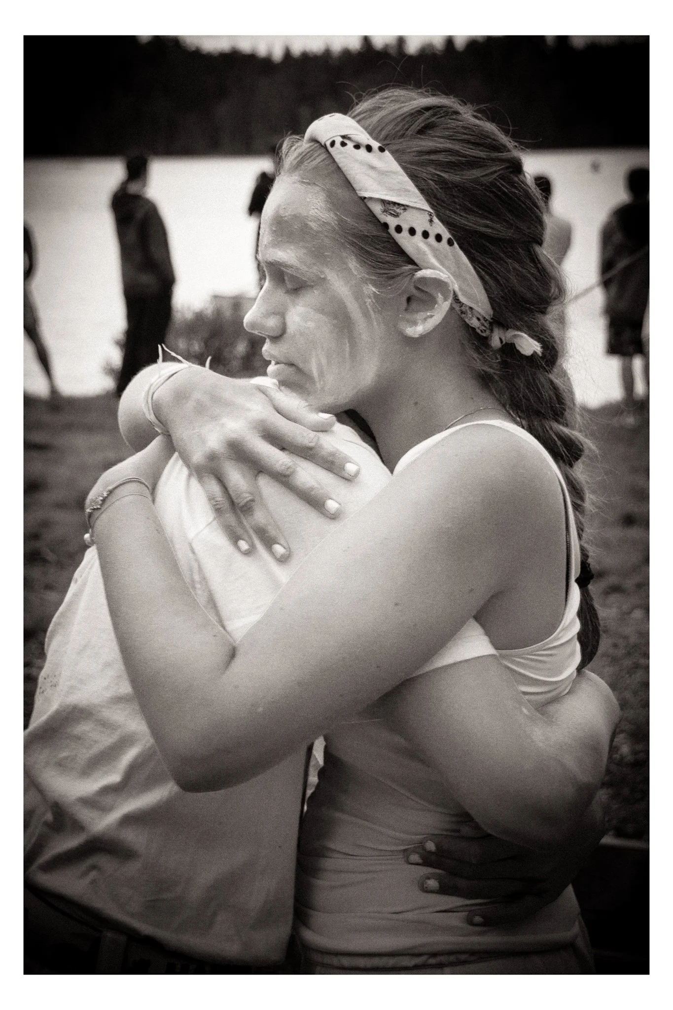 A woman and a man embrace tightly beside a lake, with trees in the background. The woman has a bandana in her hair and is wearing a tank top. The image is in black and white.