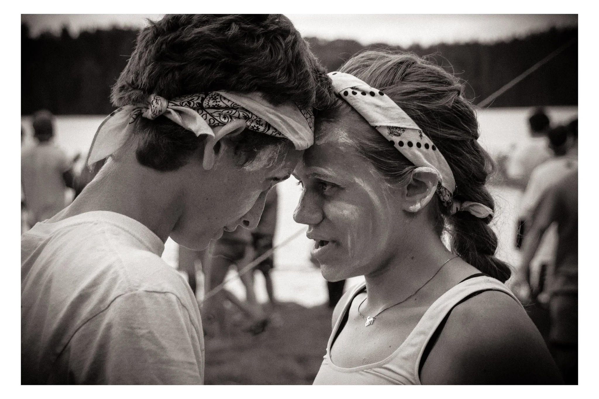 Two women with headbands touching foreheads, face paint on their faces, outdoors with people and trees in the background.