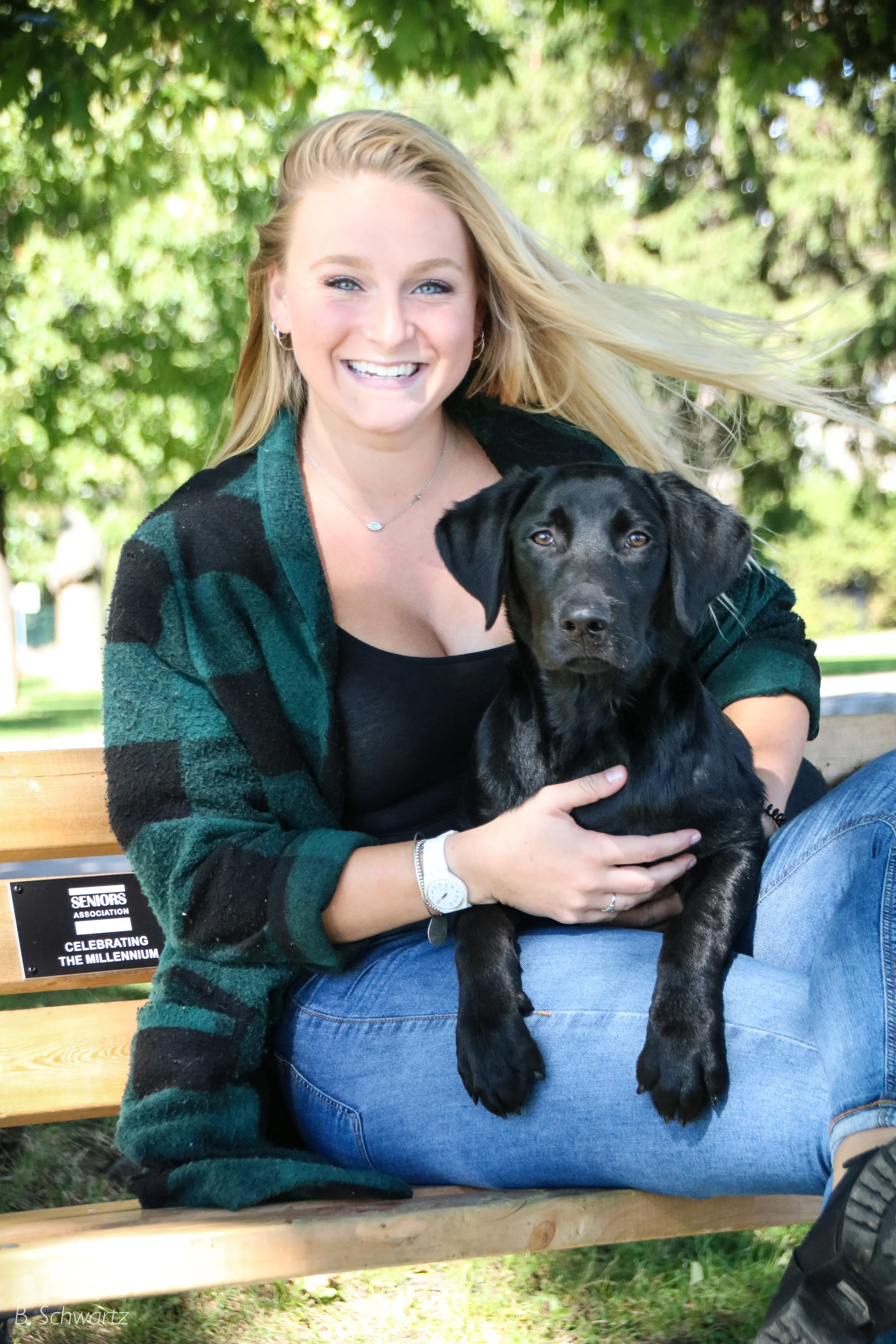 A smiling young woman with blonde hair sitting on a park bench, holding a black dog on her lap, surrounded by green trees.