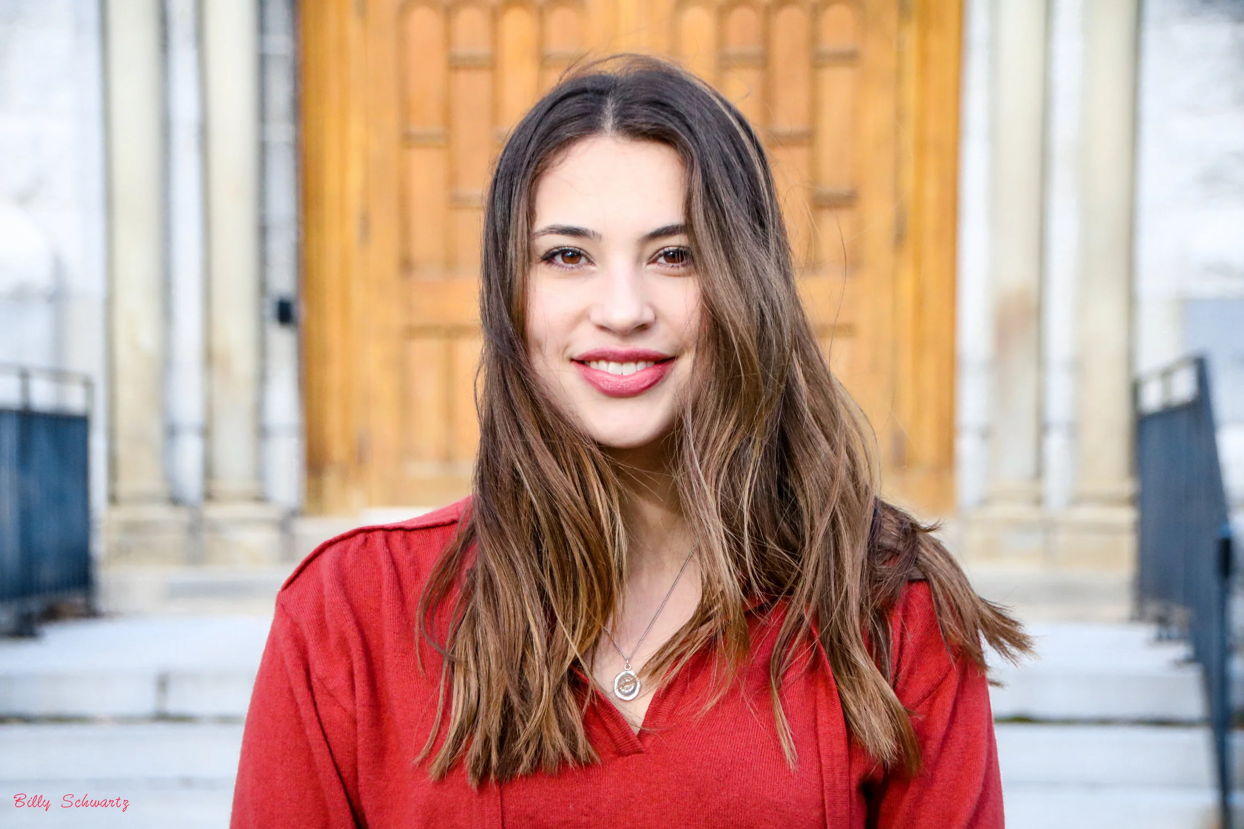 A professional corporate headshot of a young woman with long brown hair wearing a red top stands in front of steps leading to a building entrance, smiling at the camera.