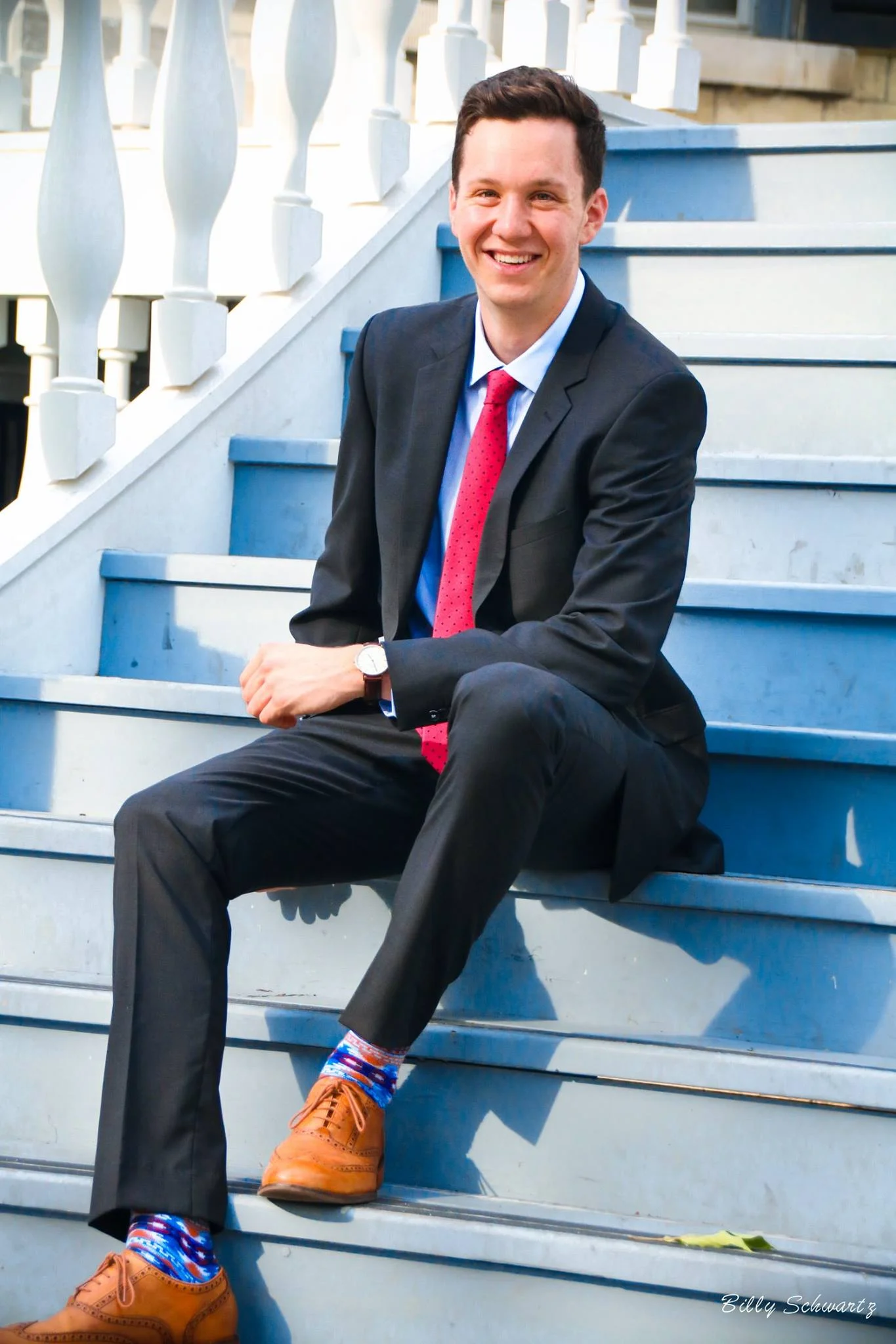 A young man in a dark suit, white shirt, red polka dot tie, brown dress shoes, and patriotic socks is sitting on outdoor blue stairs, smiling at the camera.