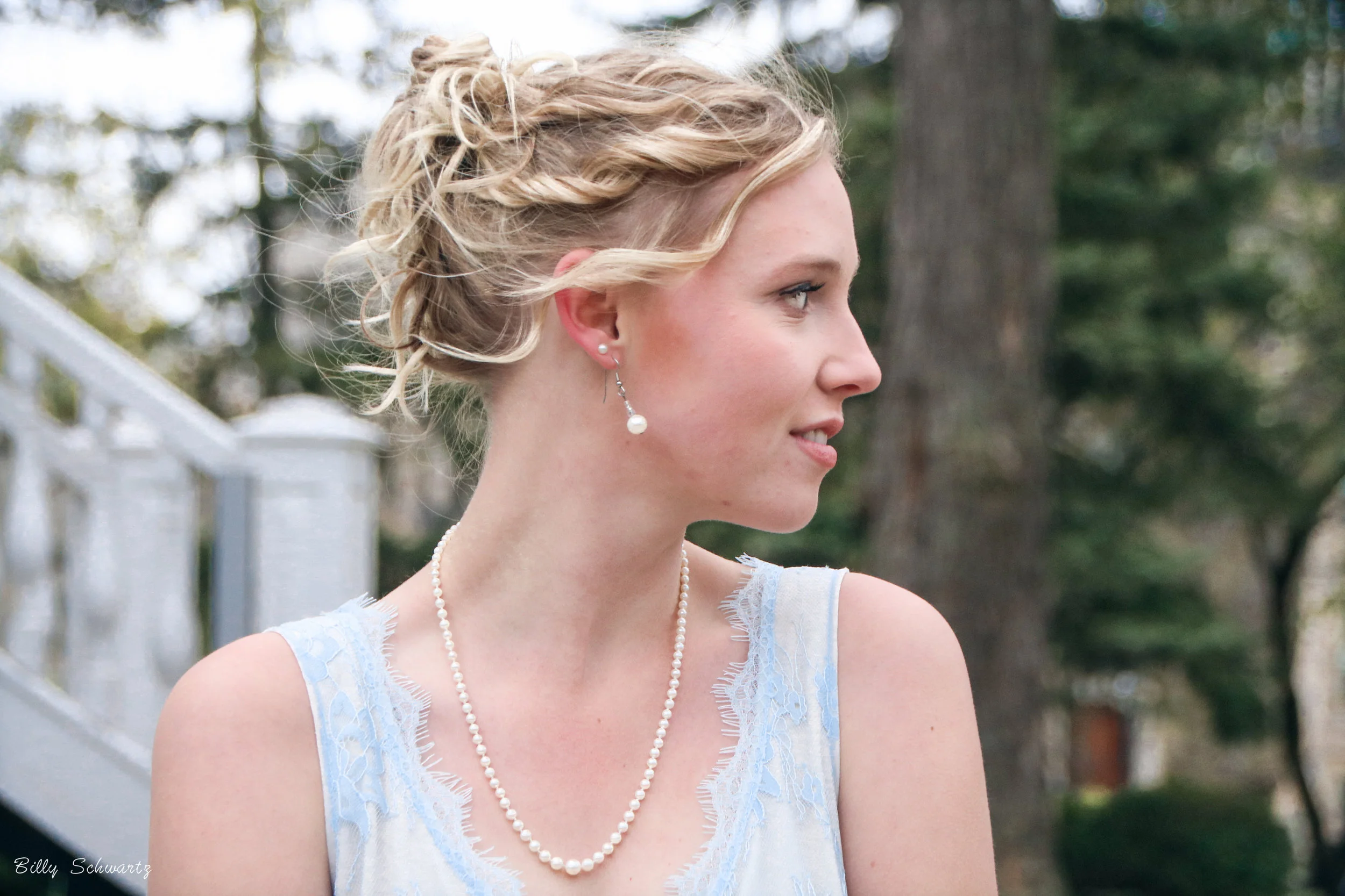 A young woman with blonde hair styled in an elegant updo wearing pearl earrings and a pearl necklace, dressed in a light-colored sleeveless dress, standing outdoors with trees and a white railing in the background.