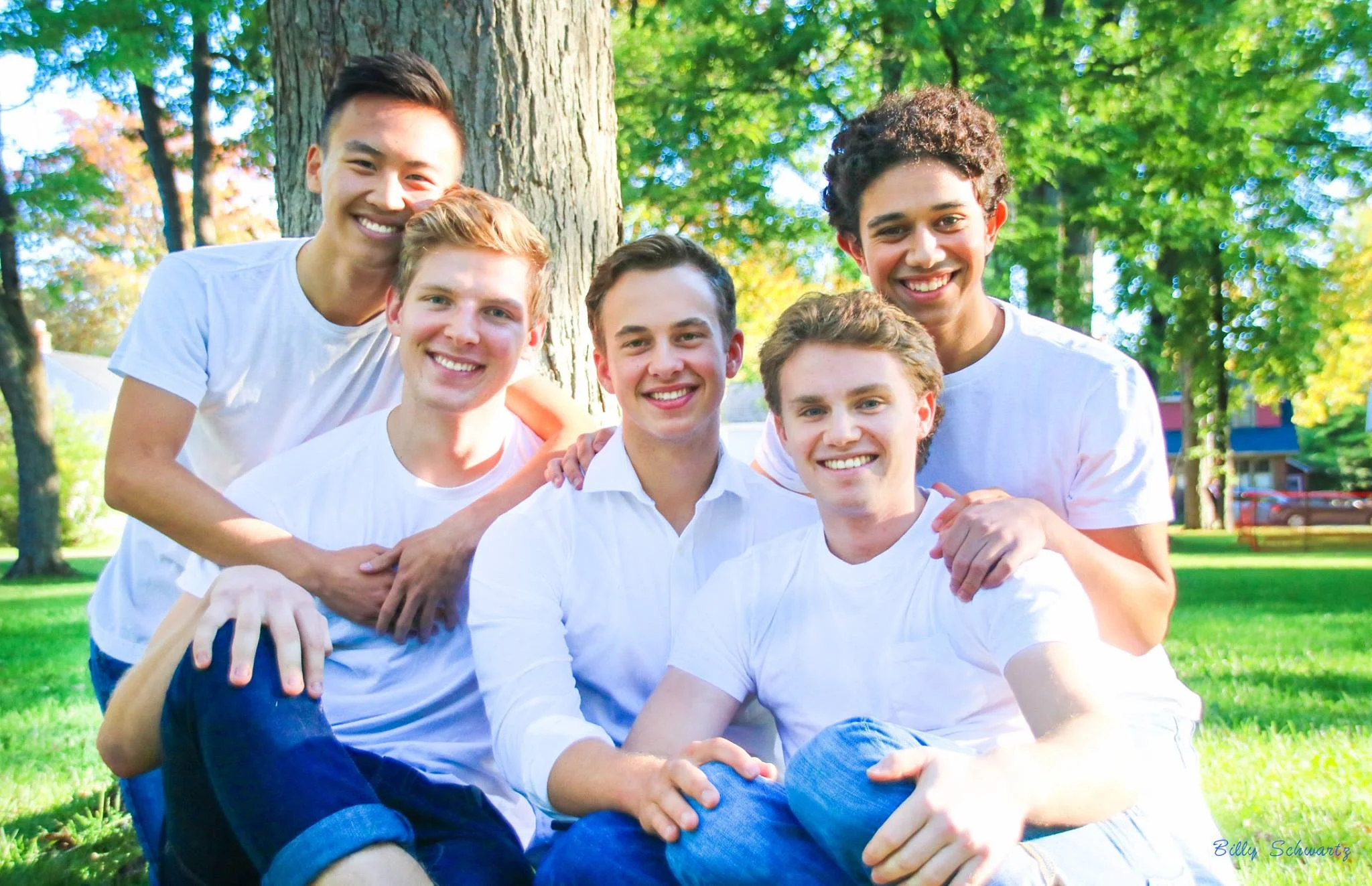Group of six young men with diverse ethnicities smiling and posing together outdoors in a park on a sunny day, with green trees and grass in the background.