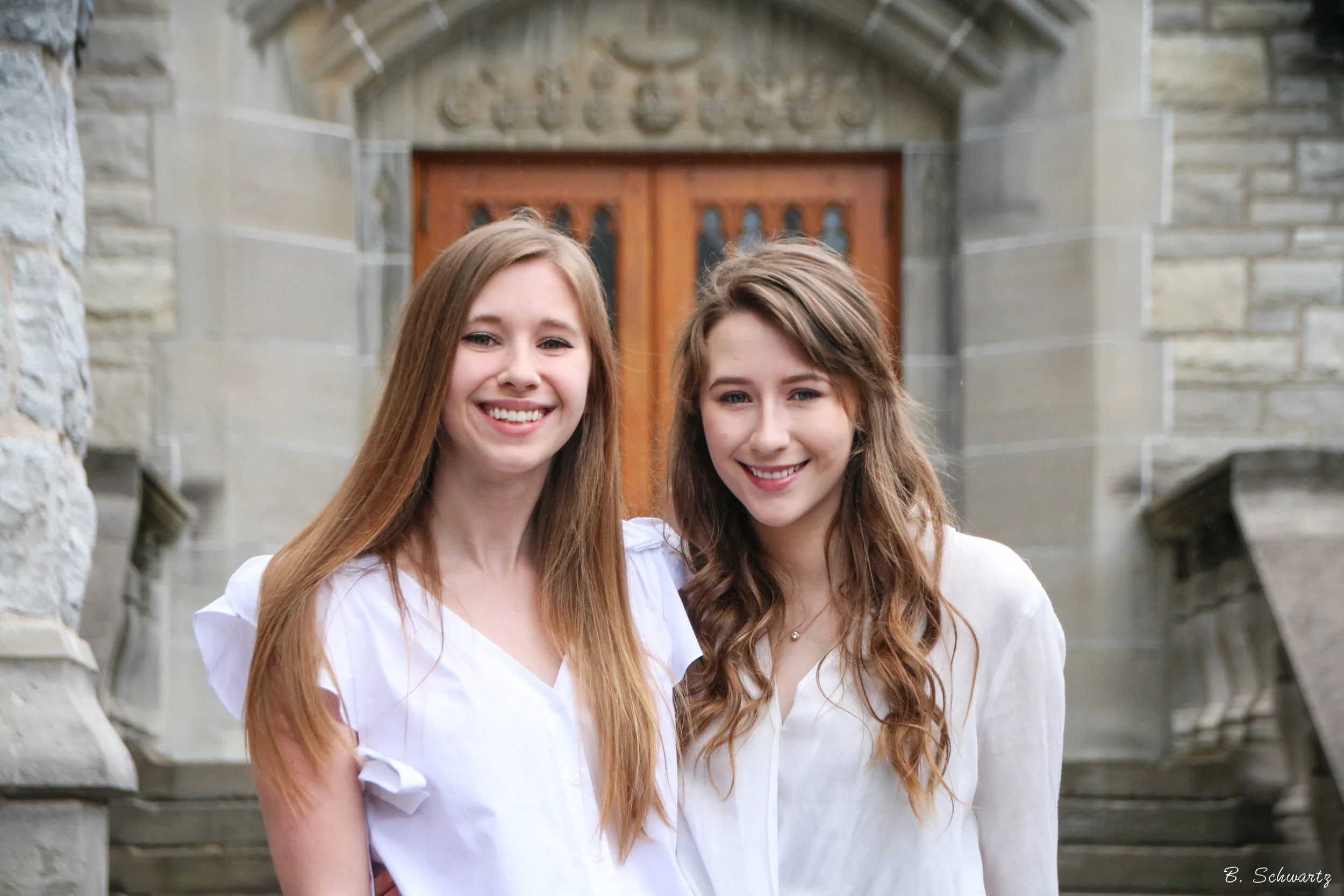 Two young women with long hair smiling outdoors in front of a stone building with a wooden door.