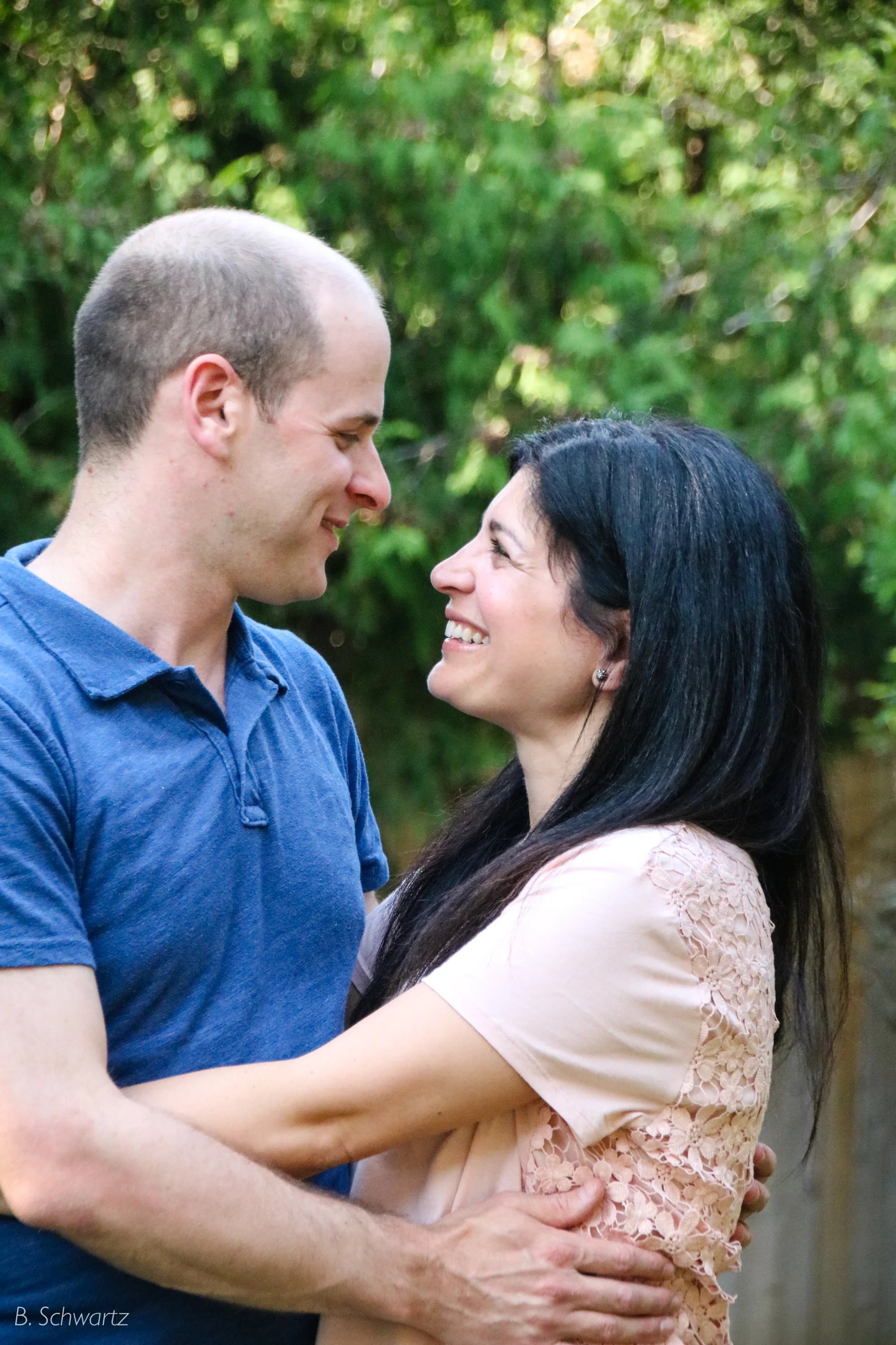 A man and woman are smiling and looking into each other's eyes outside, surrounded by green foliage.